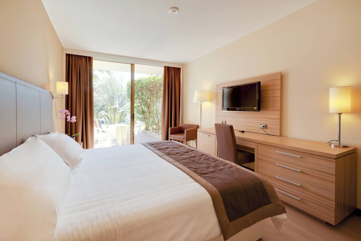Ein Hotelzimmer mit einem großen Bett, einem Schreibtisch mit Fernseher, einem Sessel und Blick ins Freie.