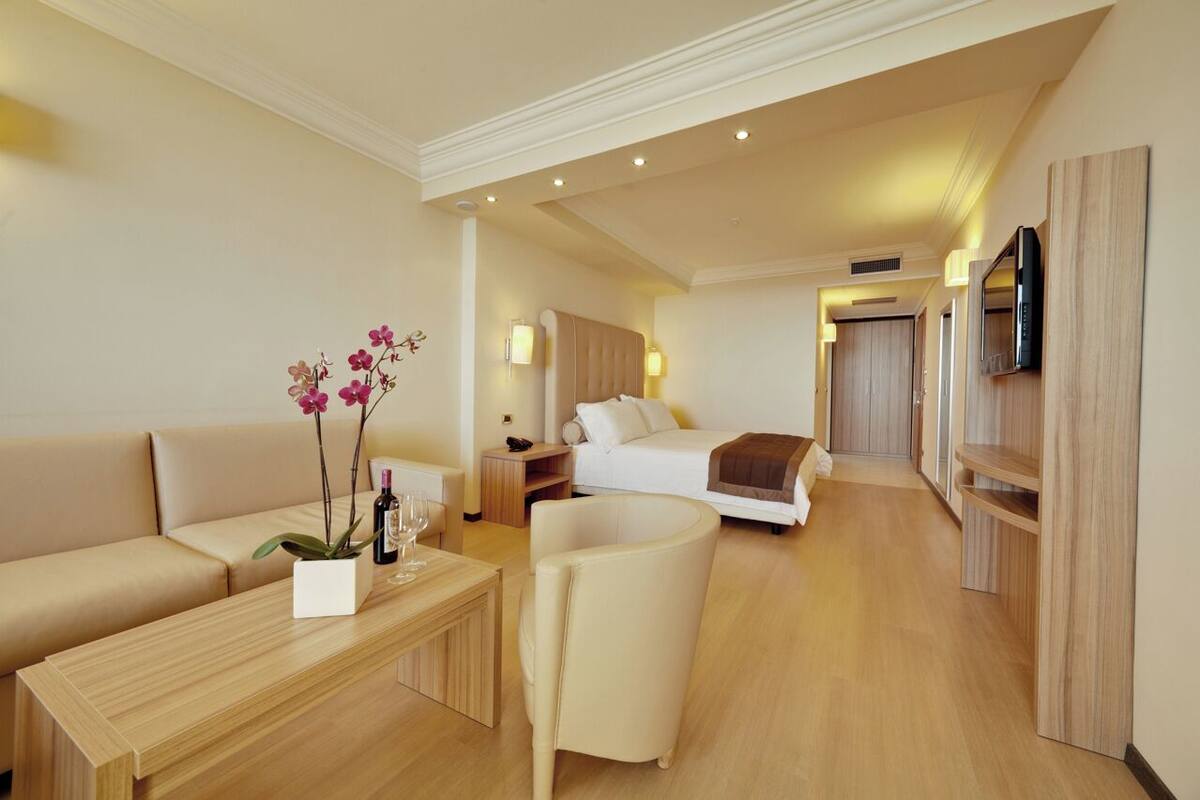 Ein modernes Hotelzimmer mit einer Couch, einem Holztisch, einem Nachttisch mit Lampe, einem Bett, einem an der Wand montierten Fernseher und einem Holzschrank.