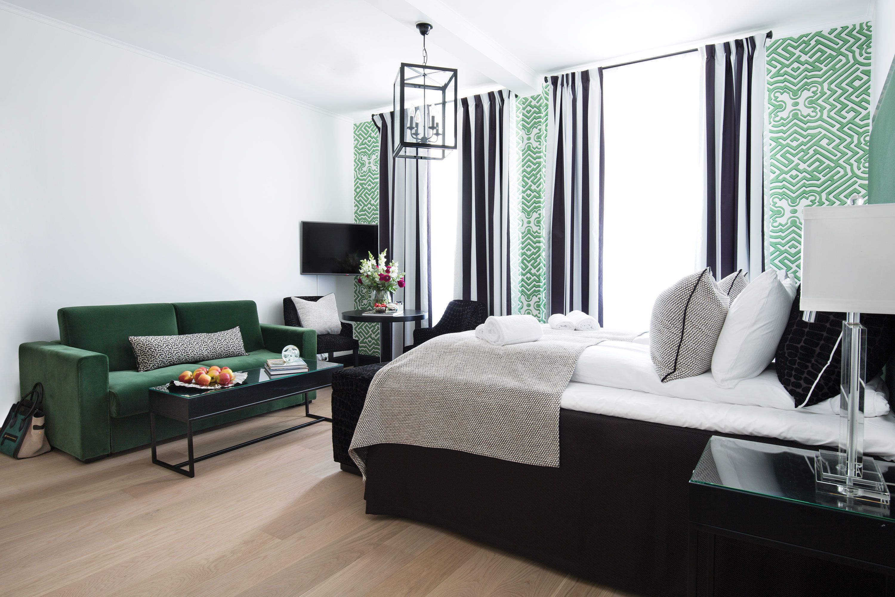Een moderne hotelkamer met een groene bank, een bed met witte beddengoed, een glazen salontafel met fruit, een nachtkastje met een lamp en gestreepte gordijnen.