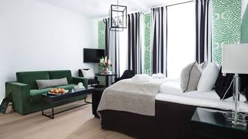 Een moderne hotelkamer met een groene bank, een bed met witte beddengoed, een glazen salontafel met fruit, een nachtkastje met een lamp en gestreepte gordijnen.