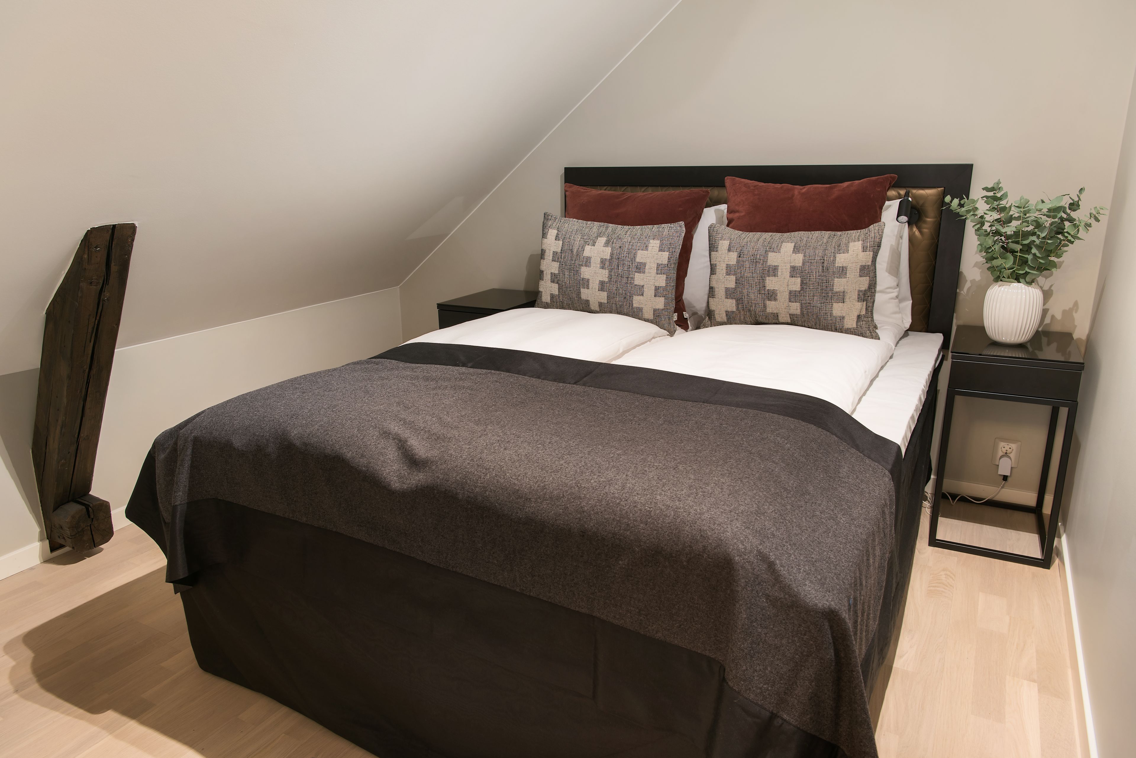 A neatly made bed with a dark bedspread and patterned pillows, a bedside table with a plant, and a wall-mounted outlet.