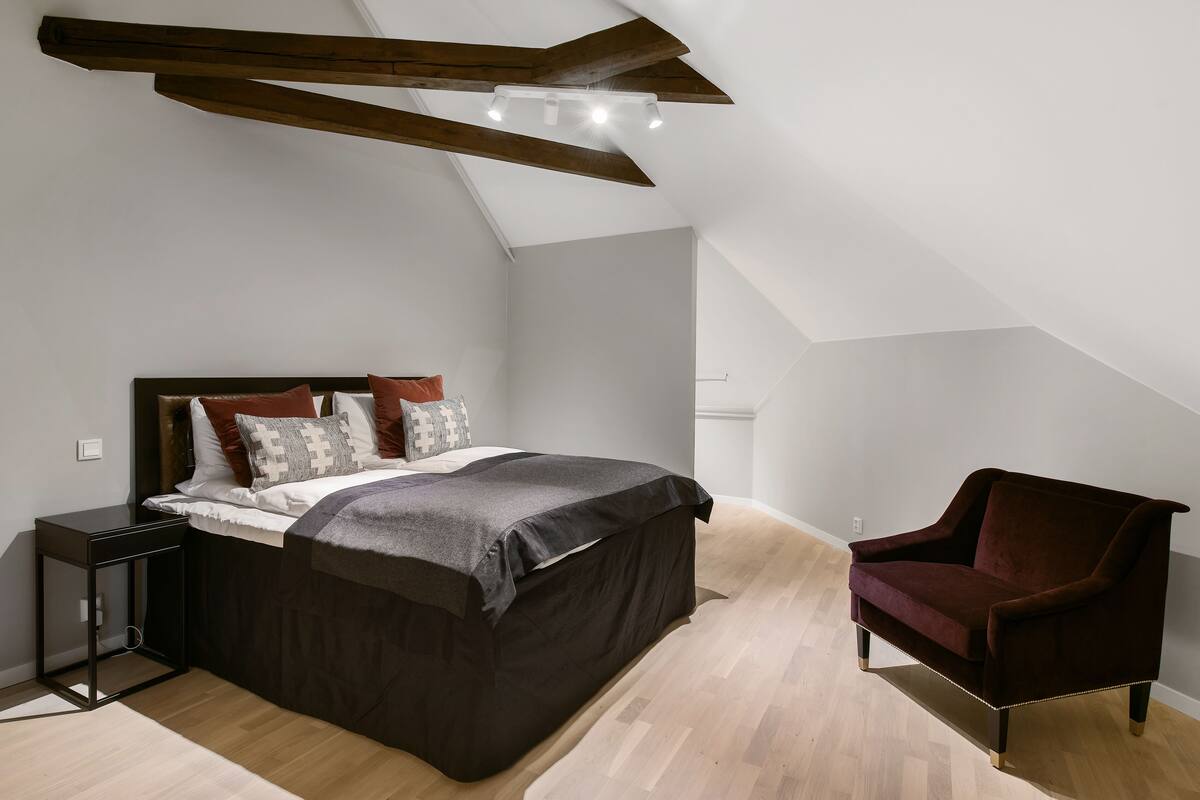 A bedroom with a bed, a nightstand, a chair, and a sloped ceiling with exposed beams.