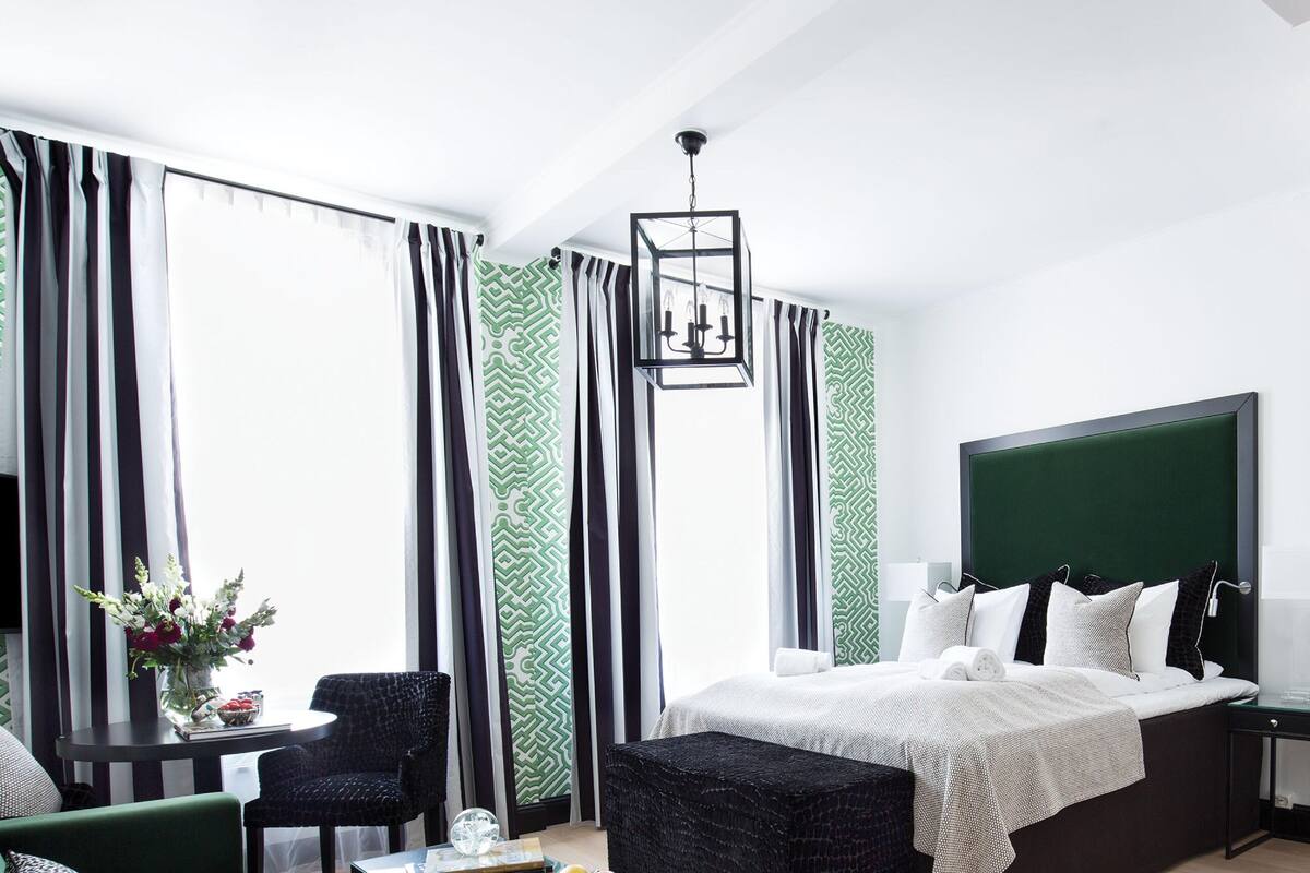 A modern hotel room with a bed, bedside table, chair, and a kitchenette area.