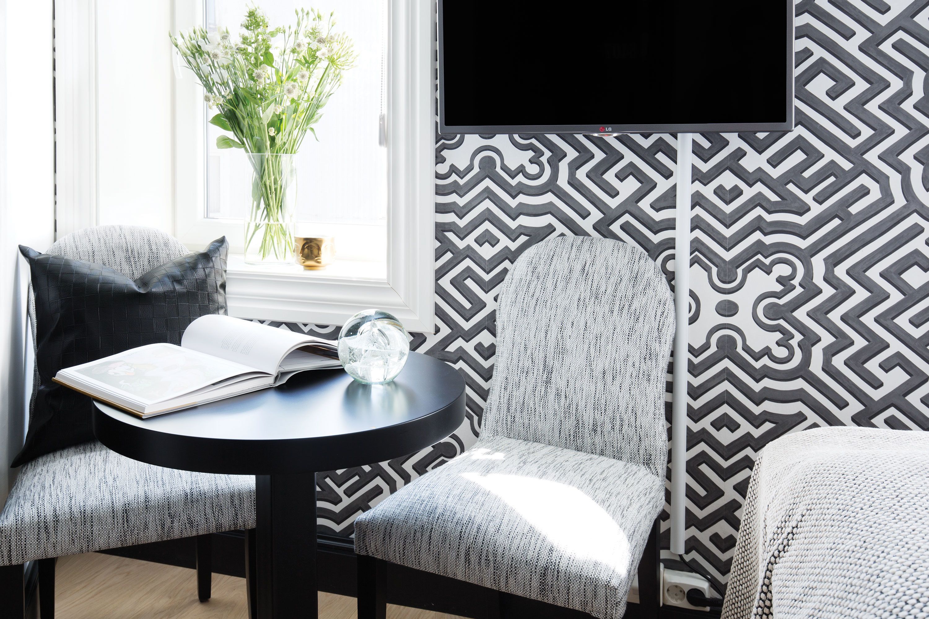 Une pièce moderne avec un mur aux motifs noir et blanc, une table ronde sur laquelle se trouve un livre et une sphère de verre, et une fenêtre agrémentée d’un vase de fleurs.