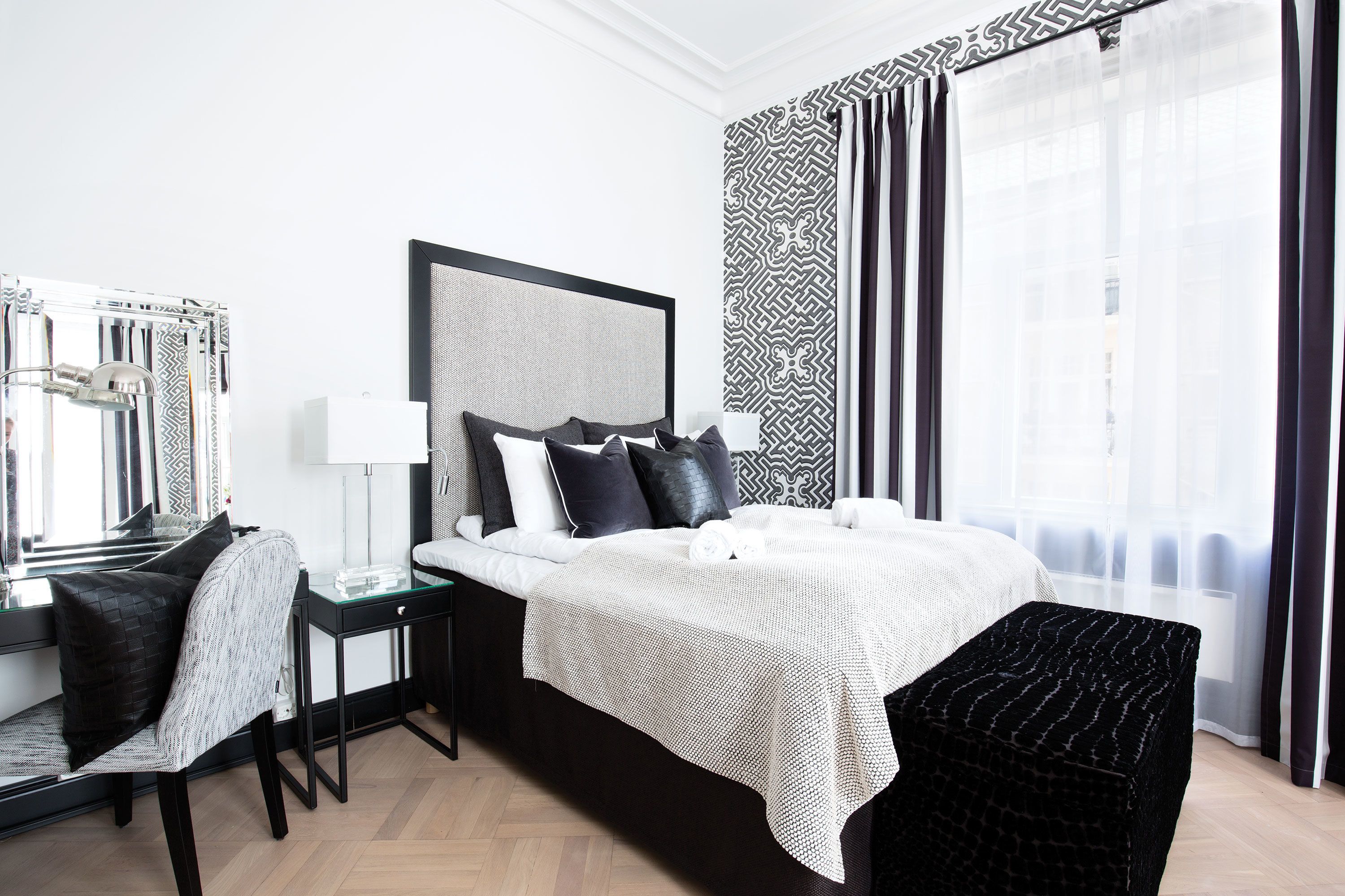 A modern hotel room with a large bed, a bedside table, a chair, and a large window with curtains.