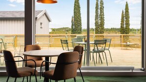 A dining area with round tables and chairs, large windows offering a view of trees and a building, and a hanging pendant light. - Scandic Gardermoen (Ullensaker)