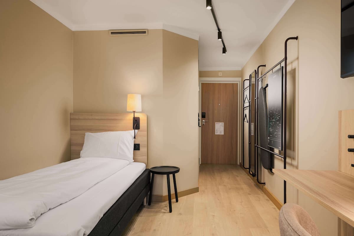 Quarto de hotel com cama, um banquinho pequeno, escrivaninha e televisão fixada na parede.