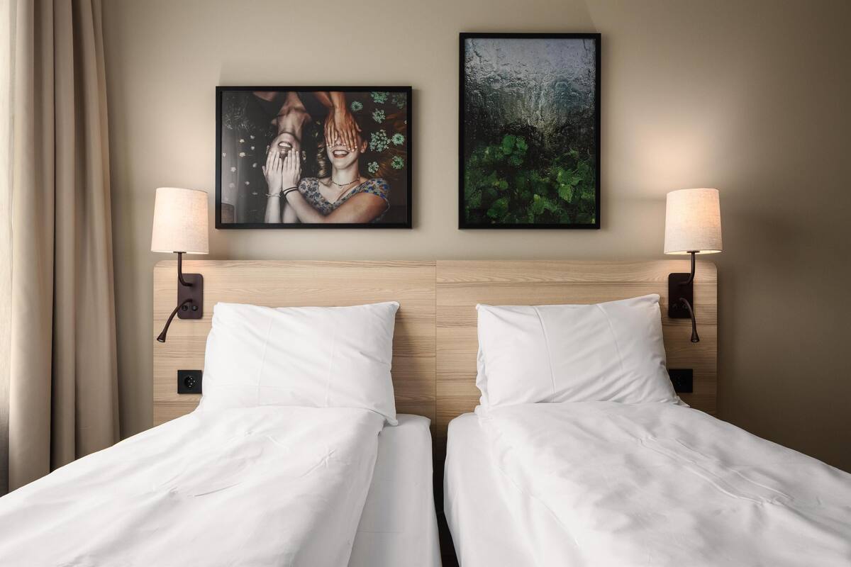 Quarto de hotel com duas camas, uma cabeceira de madeira, duas luminárias fixadas na parede e duas obras de arte emolduradas.