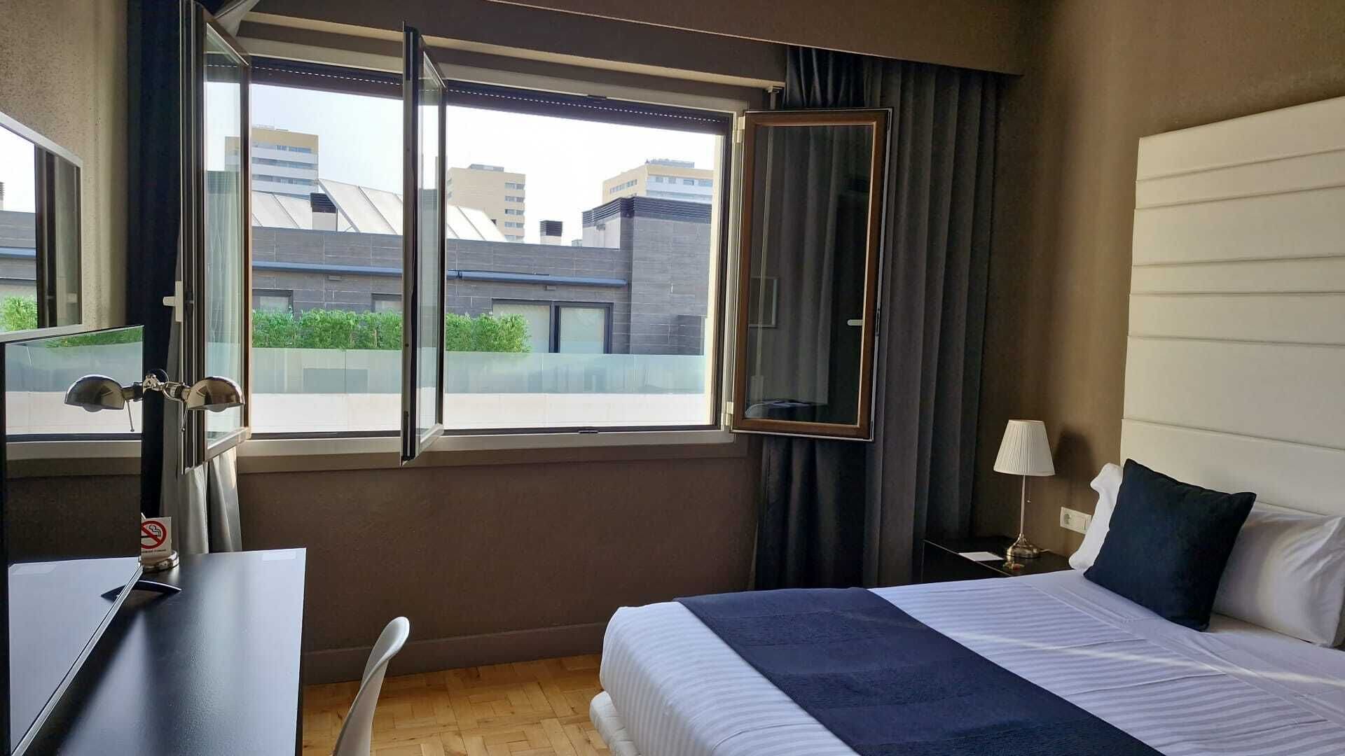 A hotel room with a bed, a desk, a chair, a window with a view of buildings, and a nightstand with a lamp.