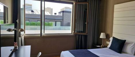 A hotel room with a bed, a desk, a chair, a window with a view of buildings, and a nightstand with a lamp.