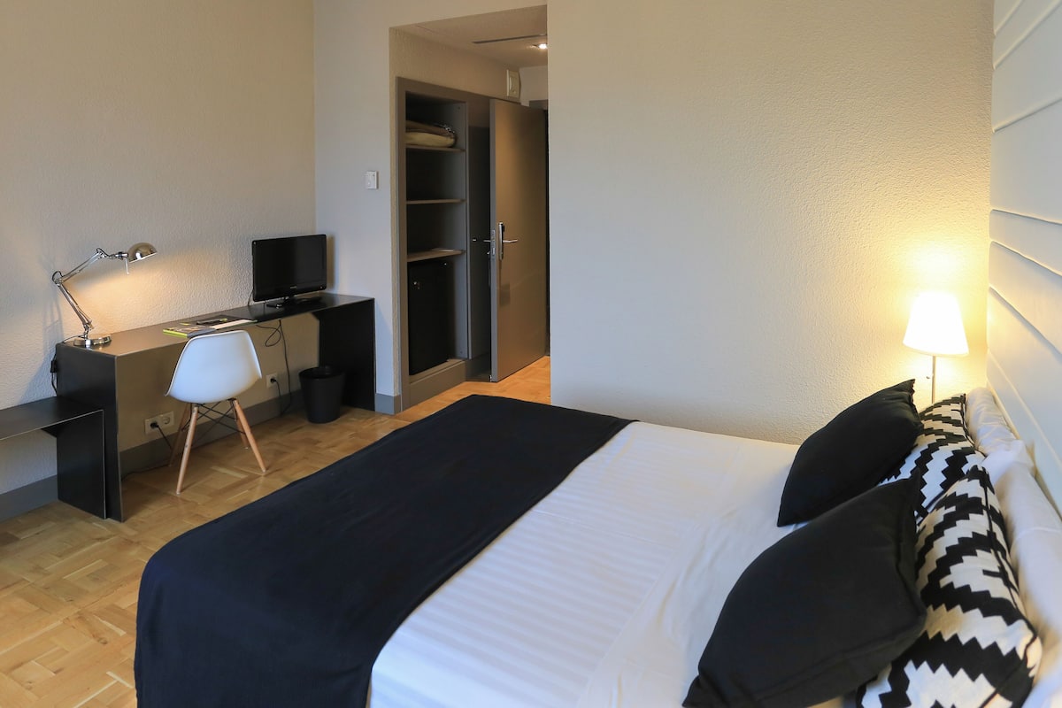 Um quarto com uma cama, uma escrivaninha com computador, uma cadeira e um guarda-roupa.