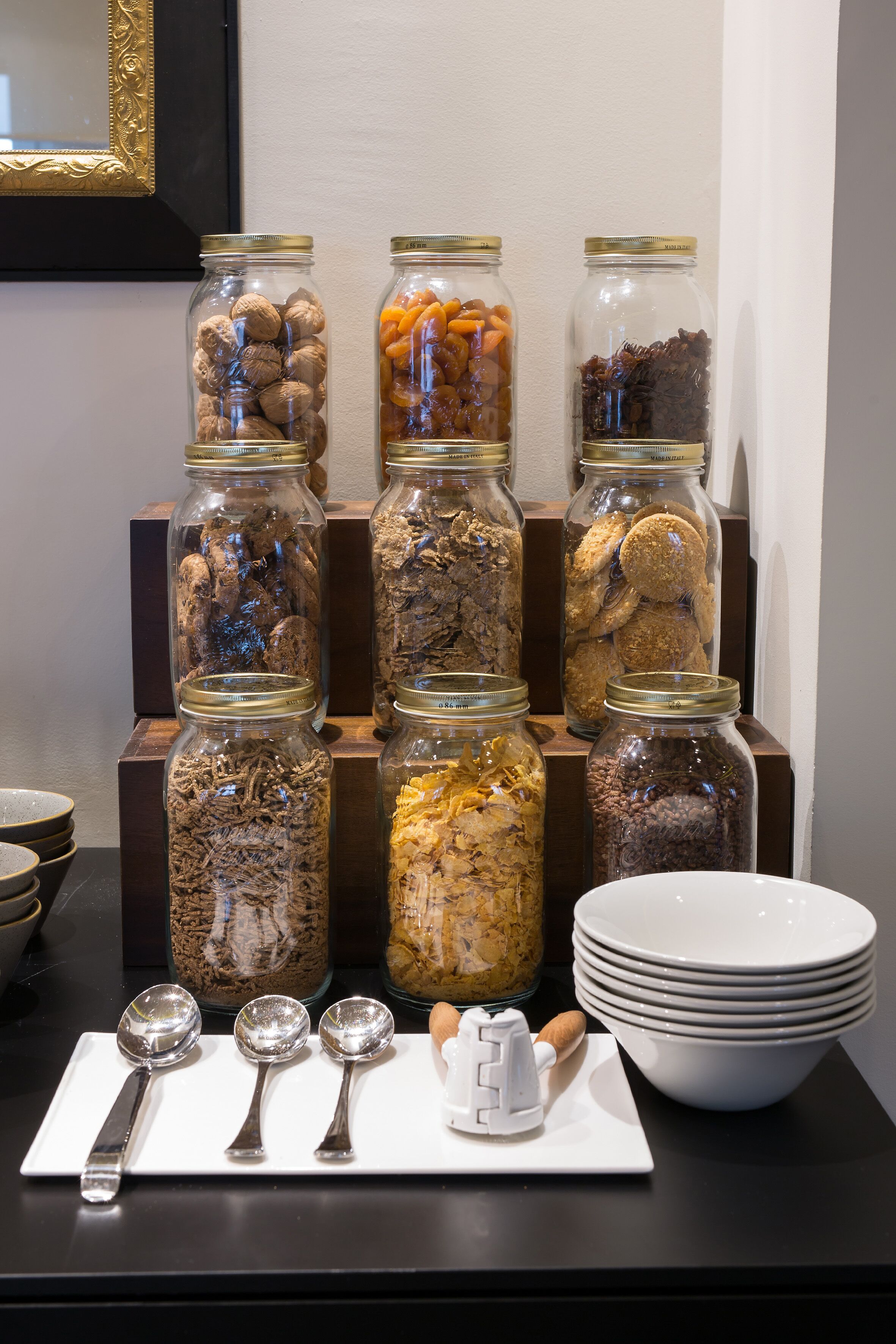 A buffet setup with jars of nuts and dried fruits, serving utensils, and a bowl of white plates.