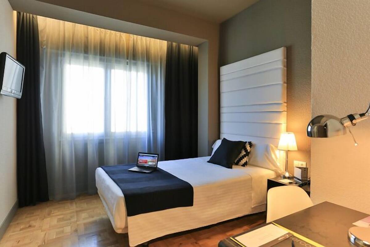 Quarto de hotel com cama, escrivaninha, cadeira, televisão na parede e janela com cortinas.