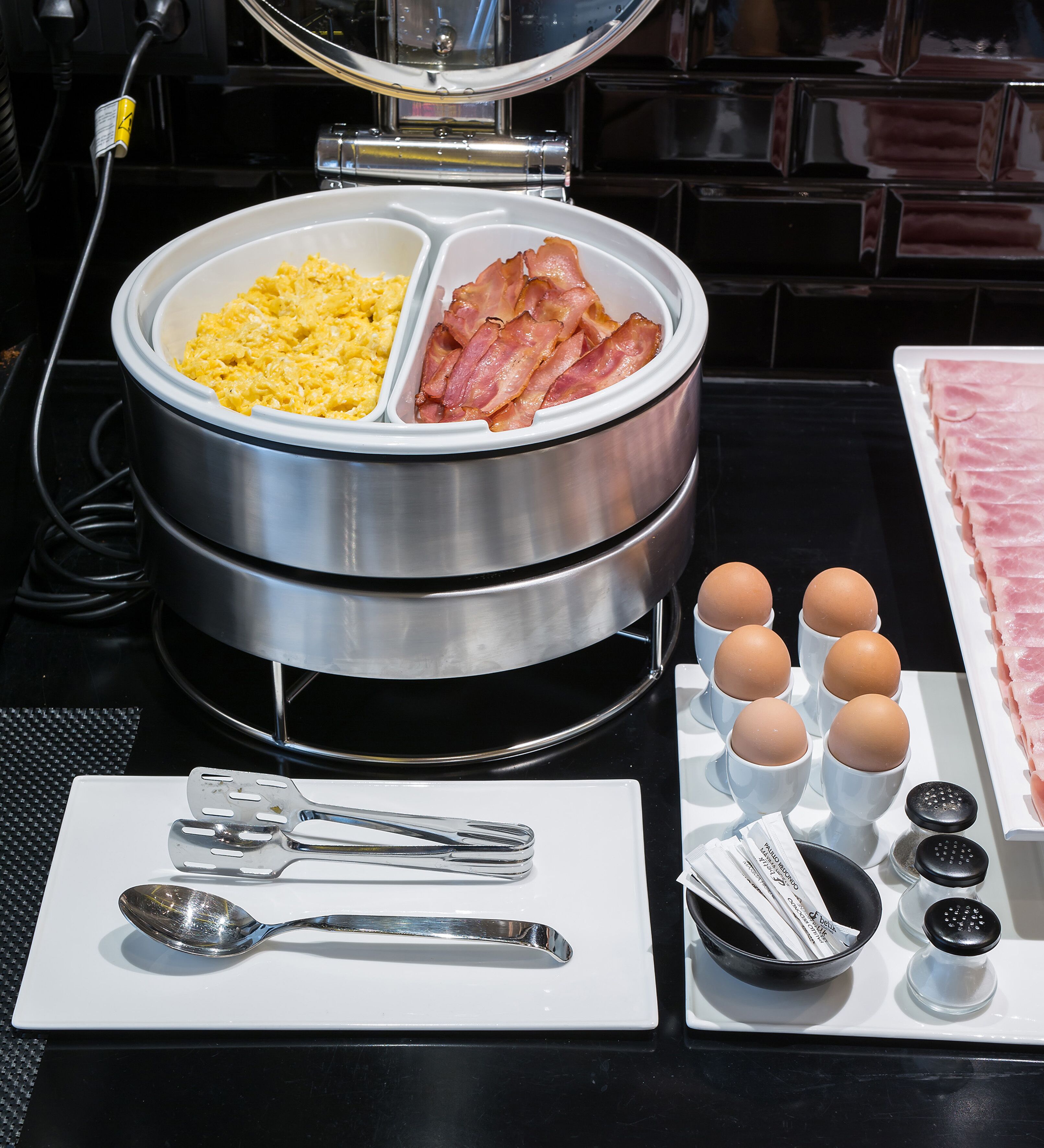 Un plateau de petit-déjeuner comprenant des œufs brouillés, du bacon et des œufs servis dans des ramequins, ainsi que des condiments et des couverts disposés sur une table.