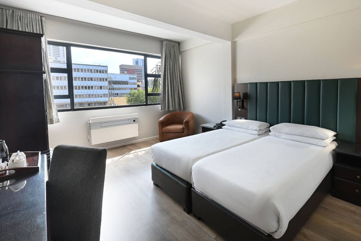 A hotel room with two beds, a desk, a chair, and a view of buildings through large windows.