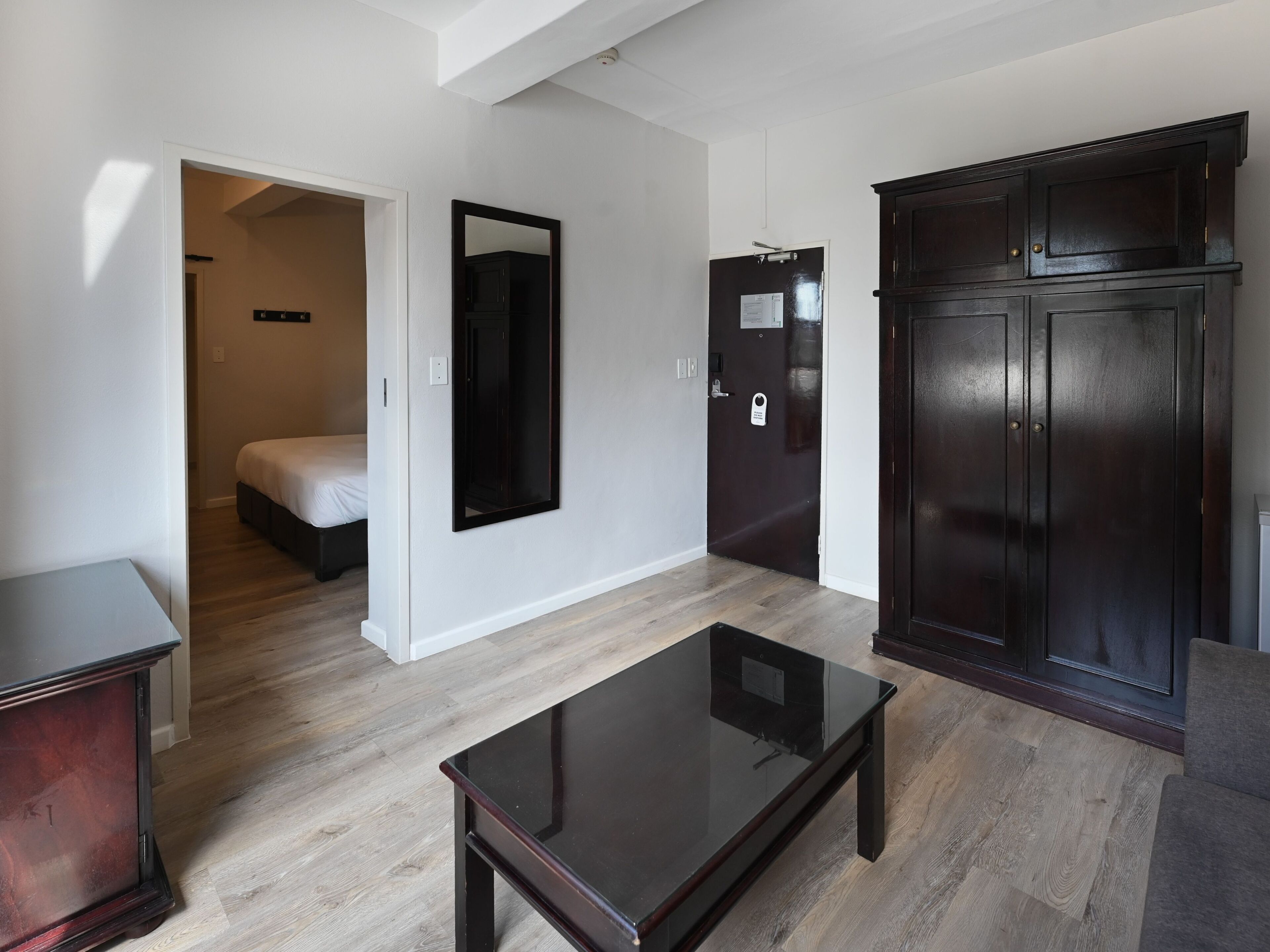 A modern room with a glass-top table, a wardrobe, a door with a lock, and a bed in the background.