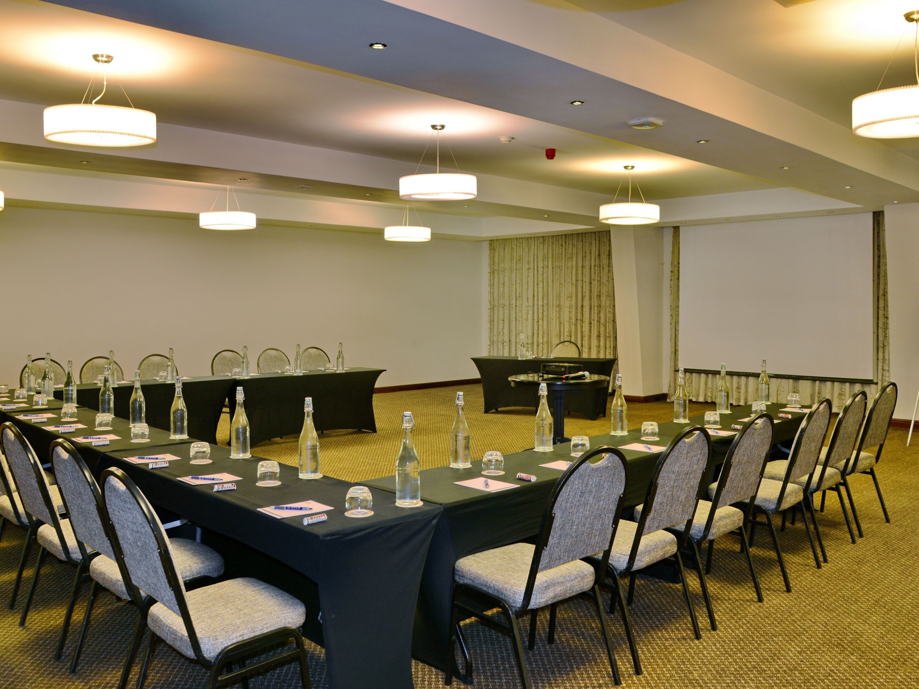 A conference room with a long table, chairs, nameplates, water bottles, and a projector screen.