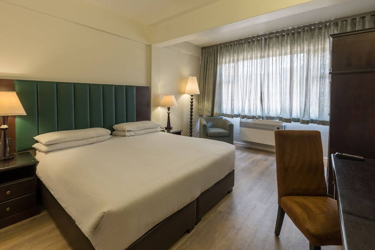 A hotel room with a large bed, two bedside lamps, a desk with a lamp, a chair, and a wardrobe.