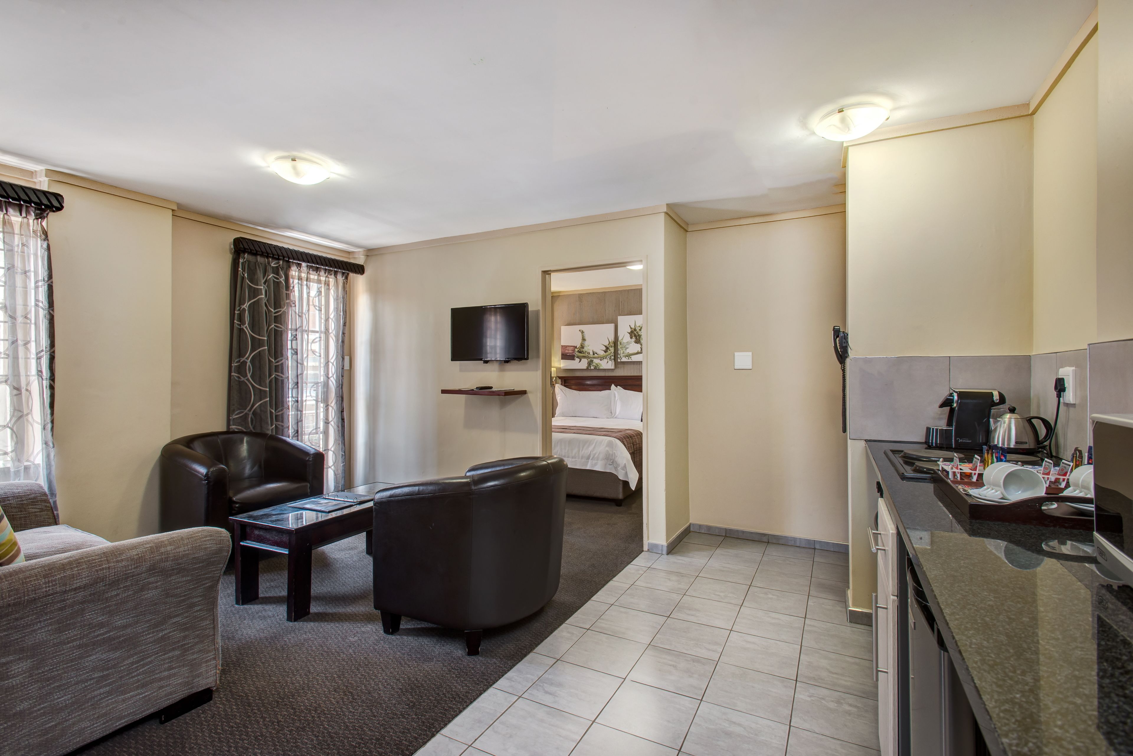 Hotelkamer met een bank, een stoel, een tafeltje, een bed, een televisie, en een keuken met koffiezetapparaat. Een badkamer is zichtbaar door een geopende deur.