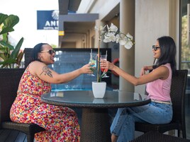 Dos personas sentadas en una mesa redonda con bebidas, una planta en maceta y un letrero al fondo.