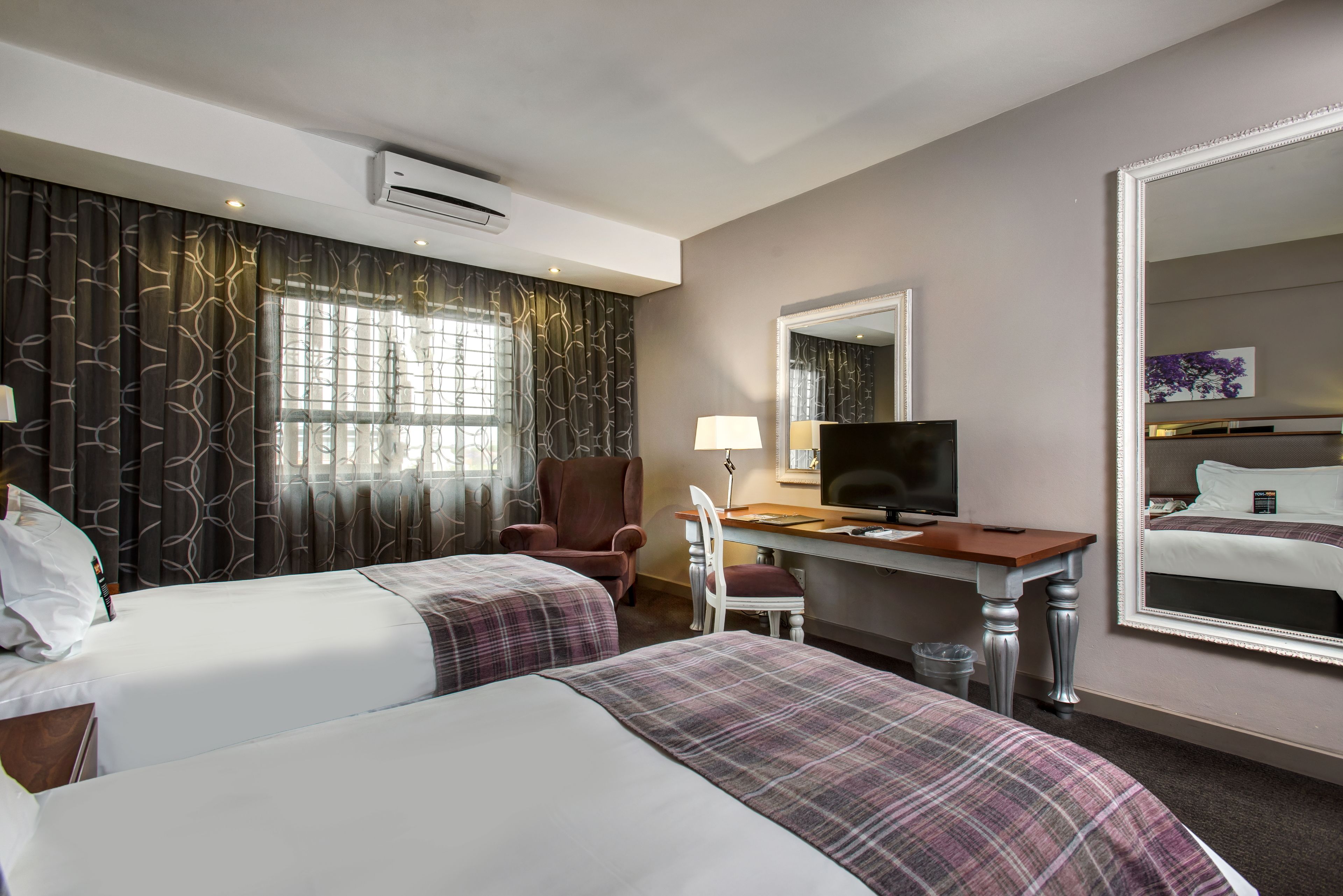 Una habitación de hotel con dos camas, un escritorio con silla, un televisor y un espejo grande.
