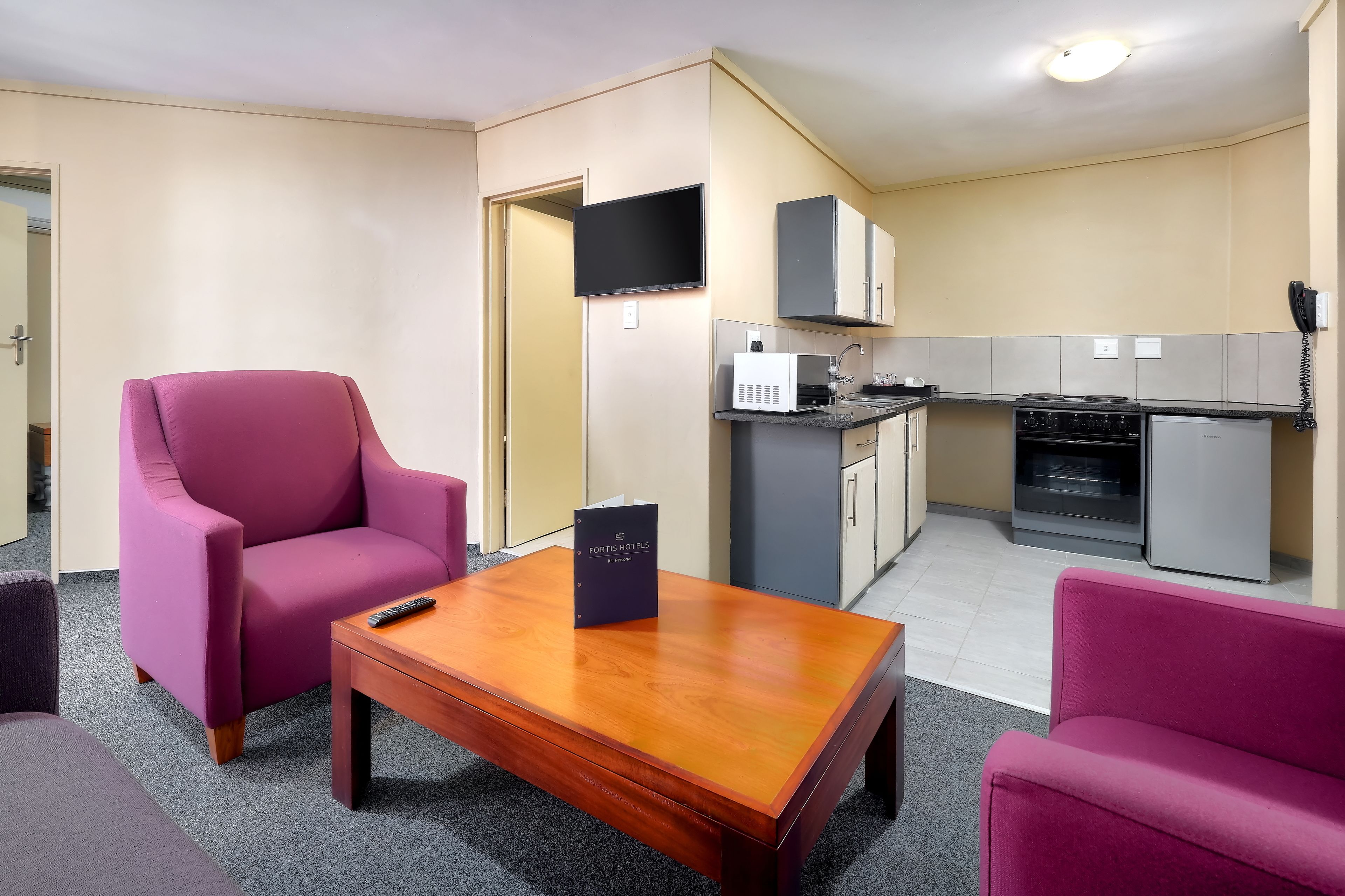 A compact hotel room with a kitchenette, a wooden coffee table, and two pink armchairs.