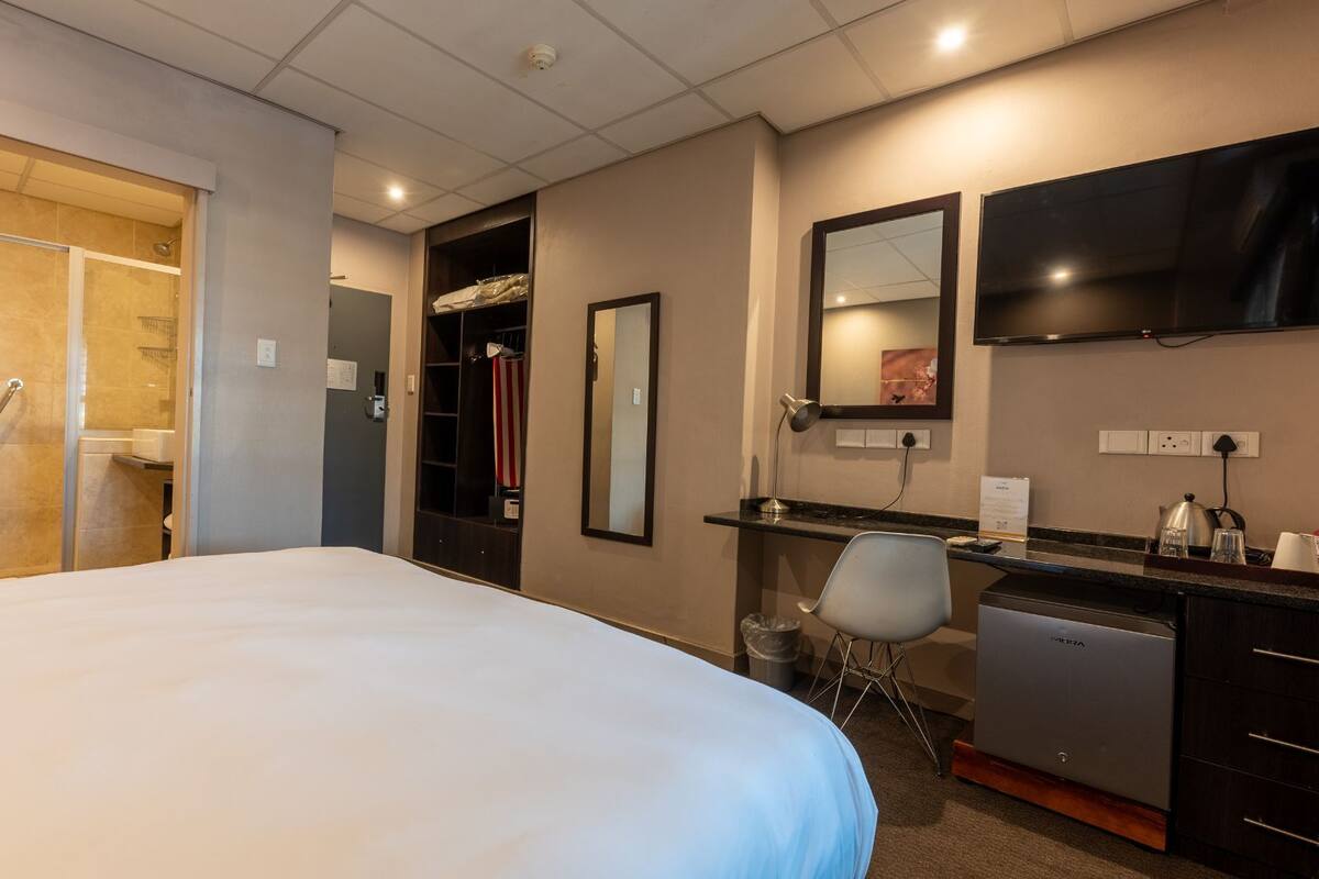 Une chambre d’hôtel avec un grand lit, un bureau avec une chaise, un miroir, une télévision et une kitchenette.