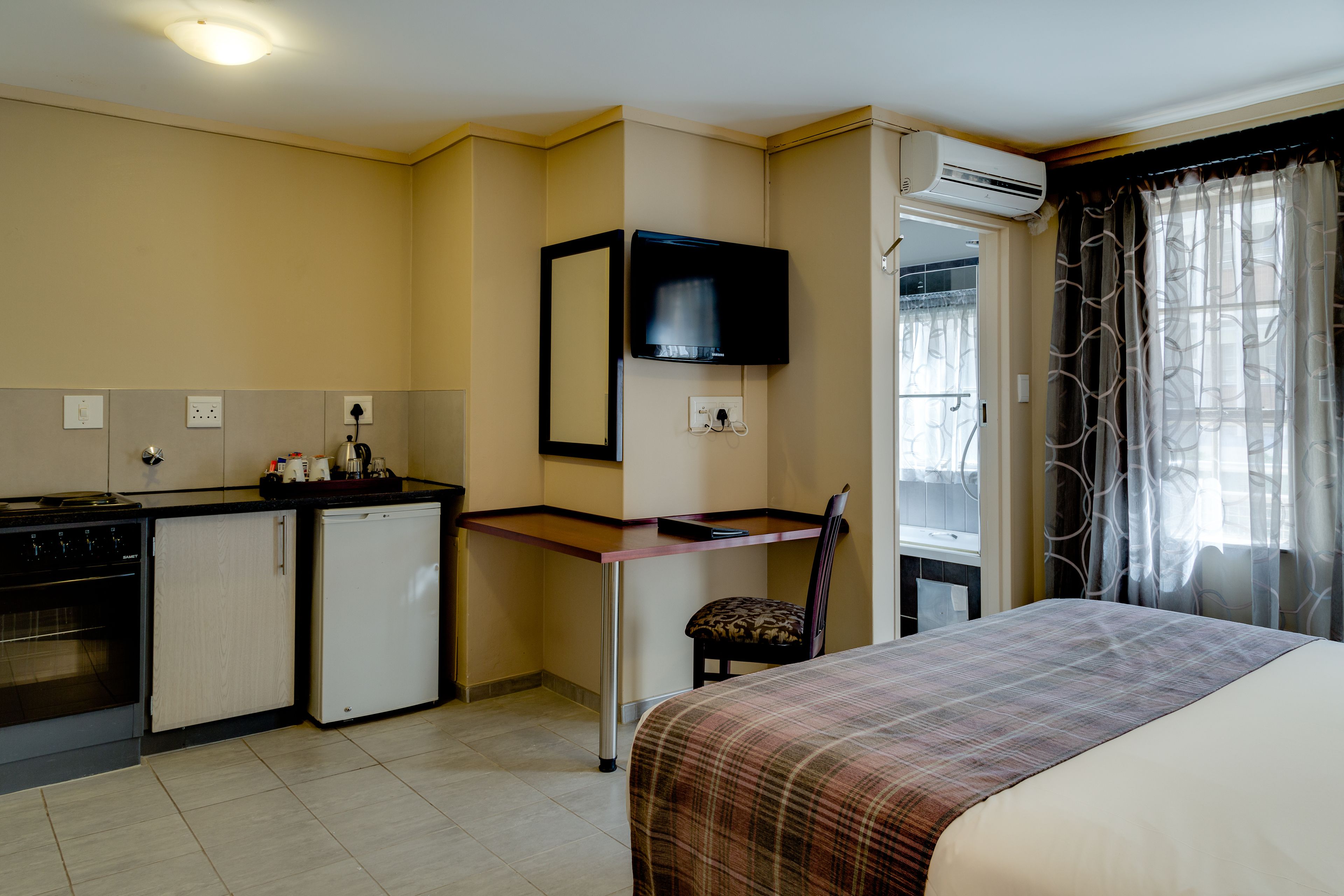 A hotel room with a bed, a desk, a chair, a kitchenette, and an air conditioning unit.