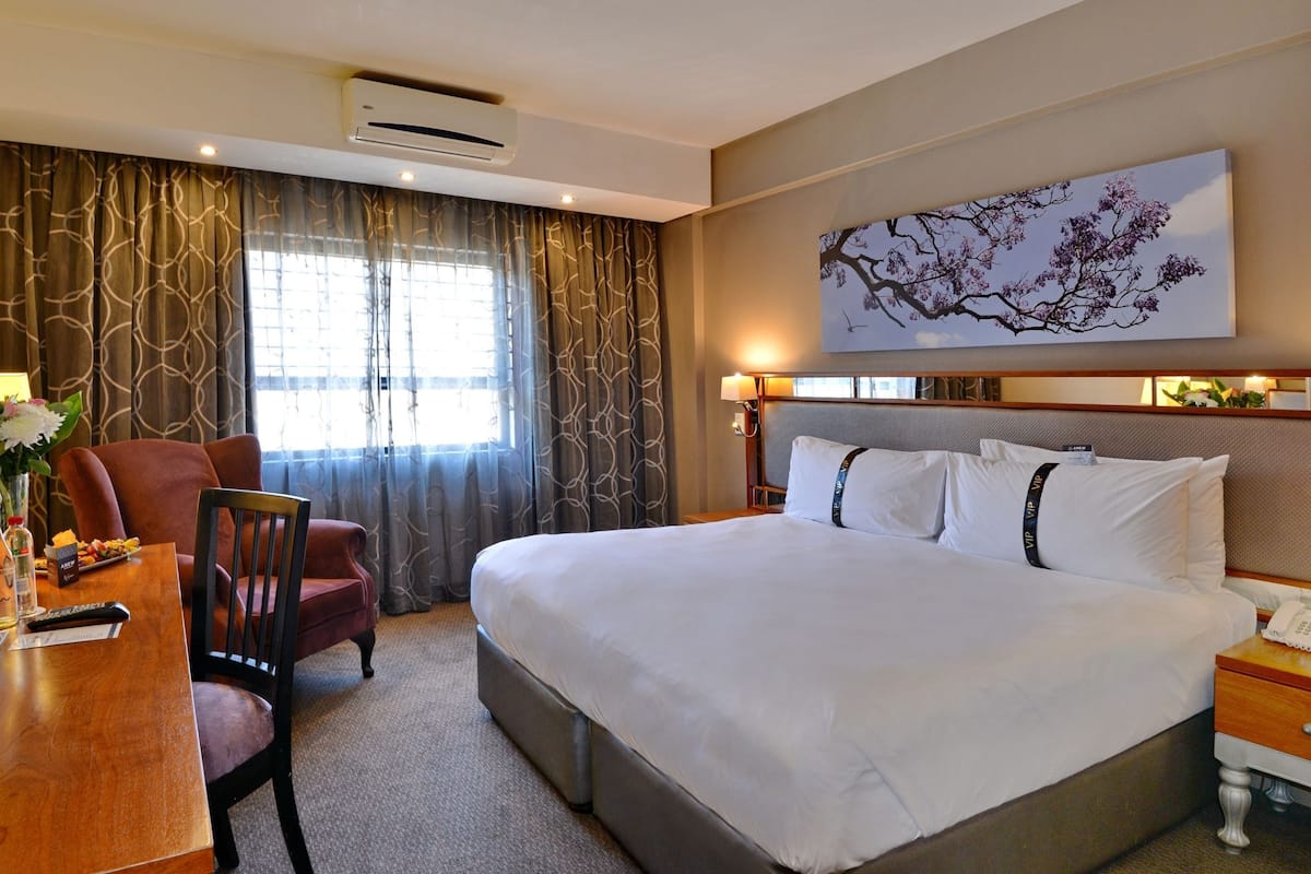 Une chambre d’hôtel avec un grand lit, un bureau avec une chaise, une table de chevet avec une lampe et une grande fenêtre avec des rideaux.