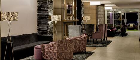 Un vestíbulo de hotel con un sofá de color oscuro, sillones estampados, un espejo grande y una chimenea.