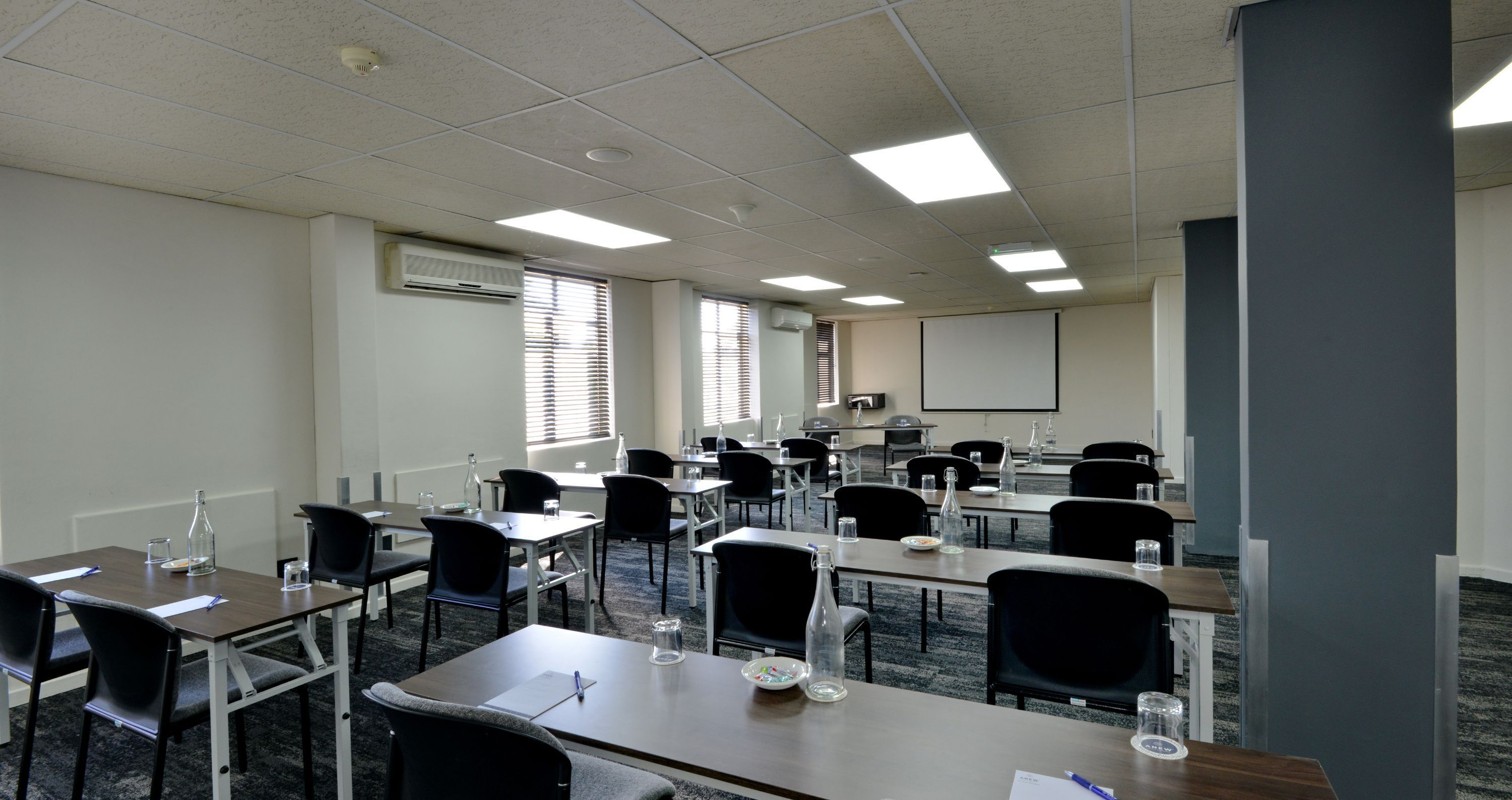 A conference room with multiple tables and chairs, equipped with water bottles and glasses, and a projector screen at the front.