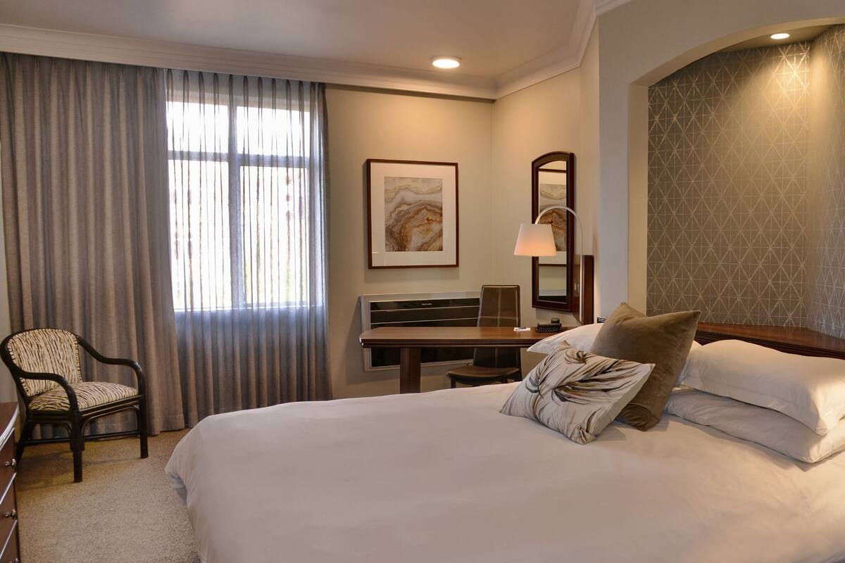 A hotel room with a large bed, a desk, a chair, a mirror, and a lamp.