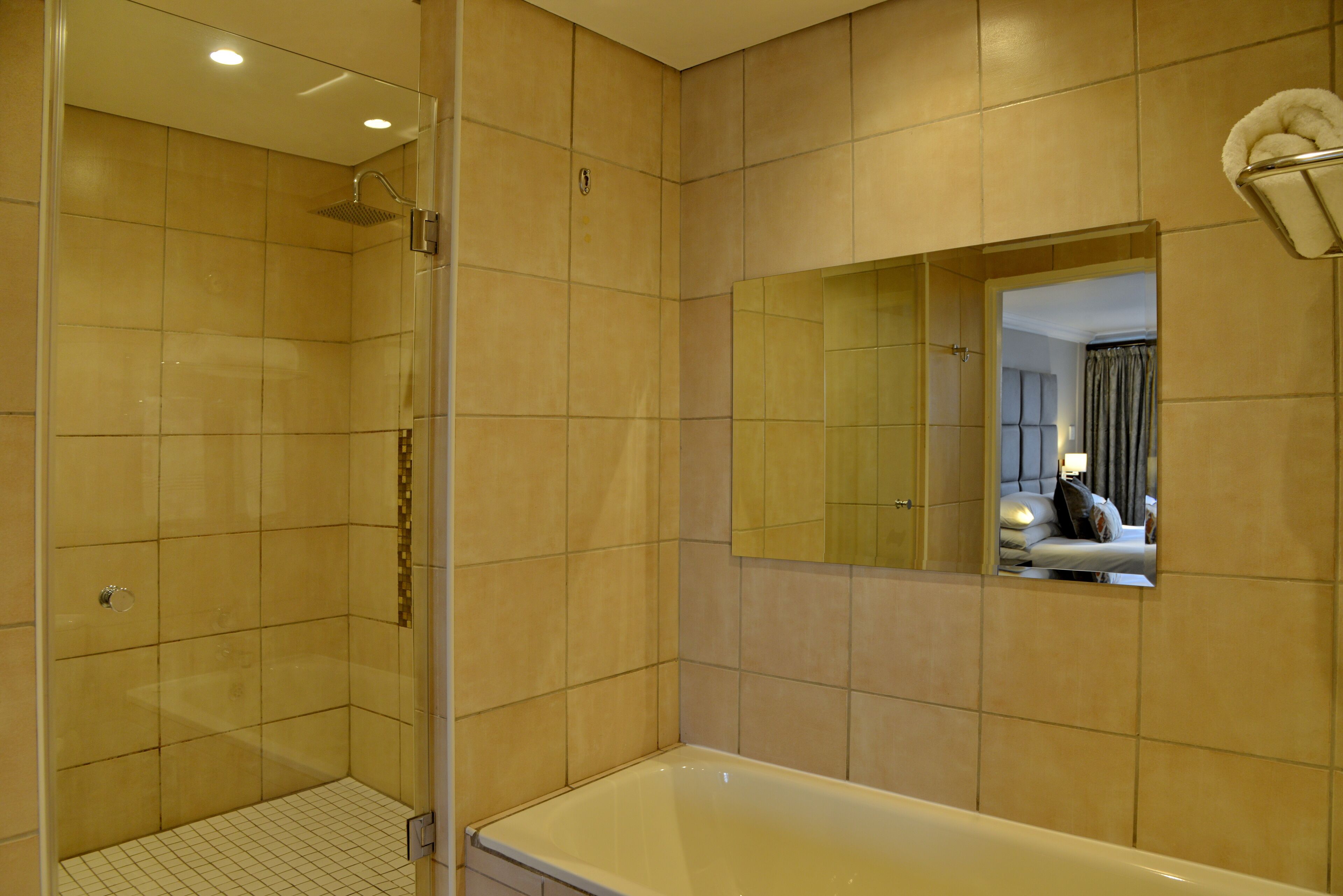 Un baño con ducha de vidrio, una bañera y un espejo que refleja un dormitorio.