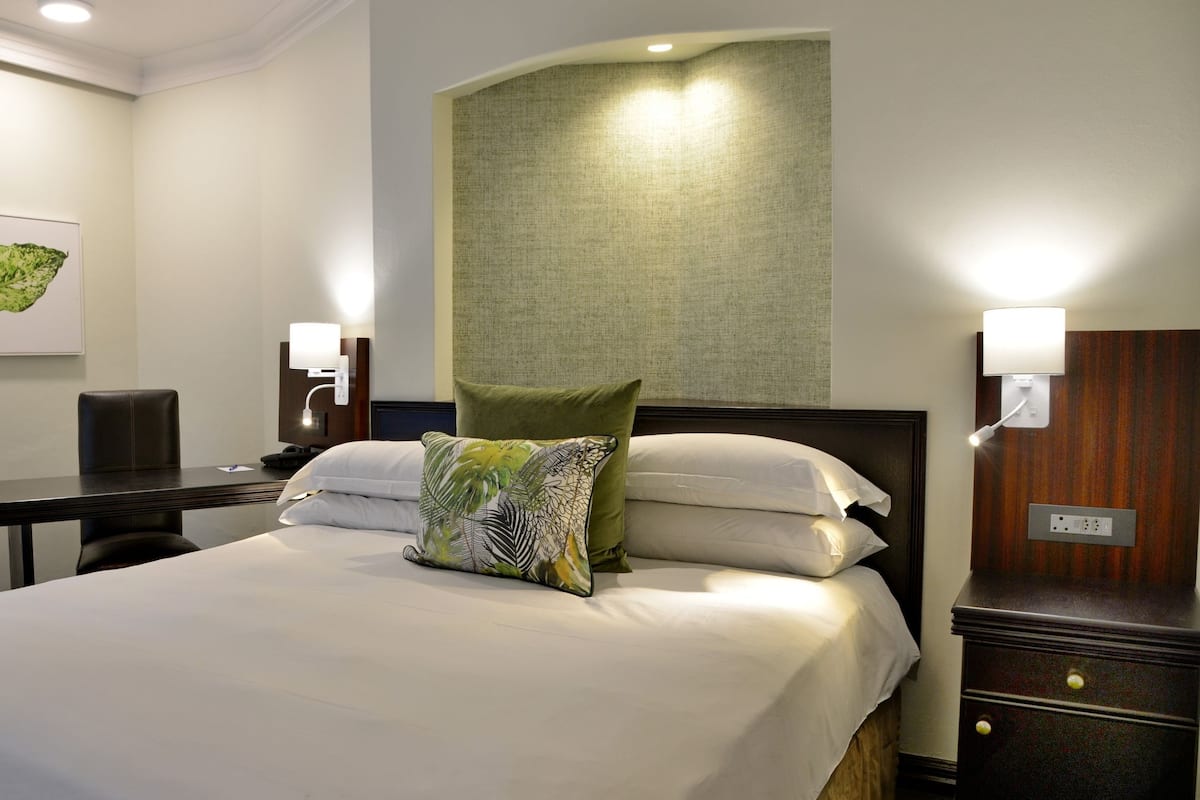 A hotel room with a bed, a desk, a chair, a lamp, and a wall-mounted light.