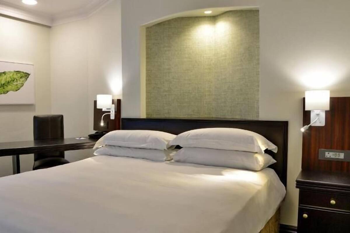 A hotel room with a bed, desk, chair, and a wall-mounted lamp.