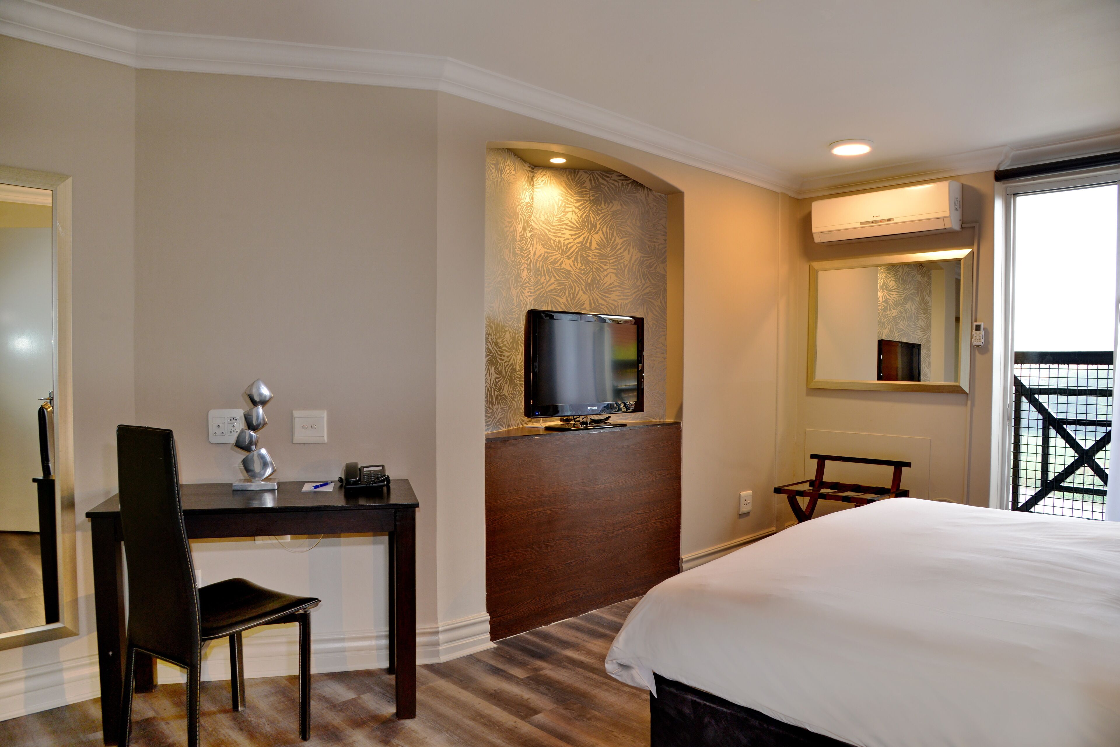 Quarto de hotel com cama, escrivaninha, cadeira, televisão e espelho.