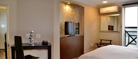 Quarto de hotel com cama, escrivaninha, cadeira, televisão e espelho.