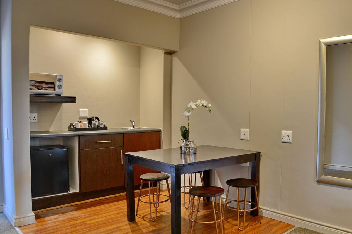 A compact kitchen with a dining table and stools, a small refrigerator, and a mirror on the wall.