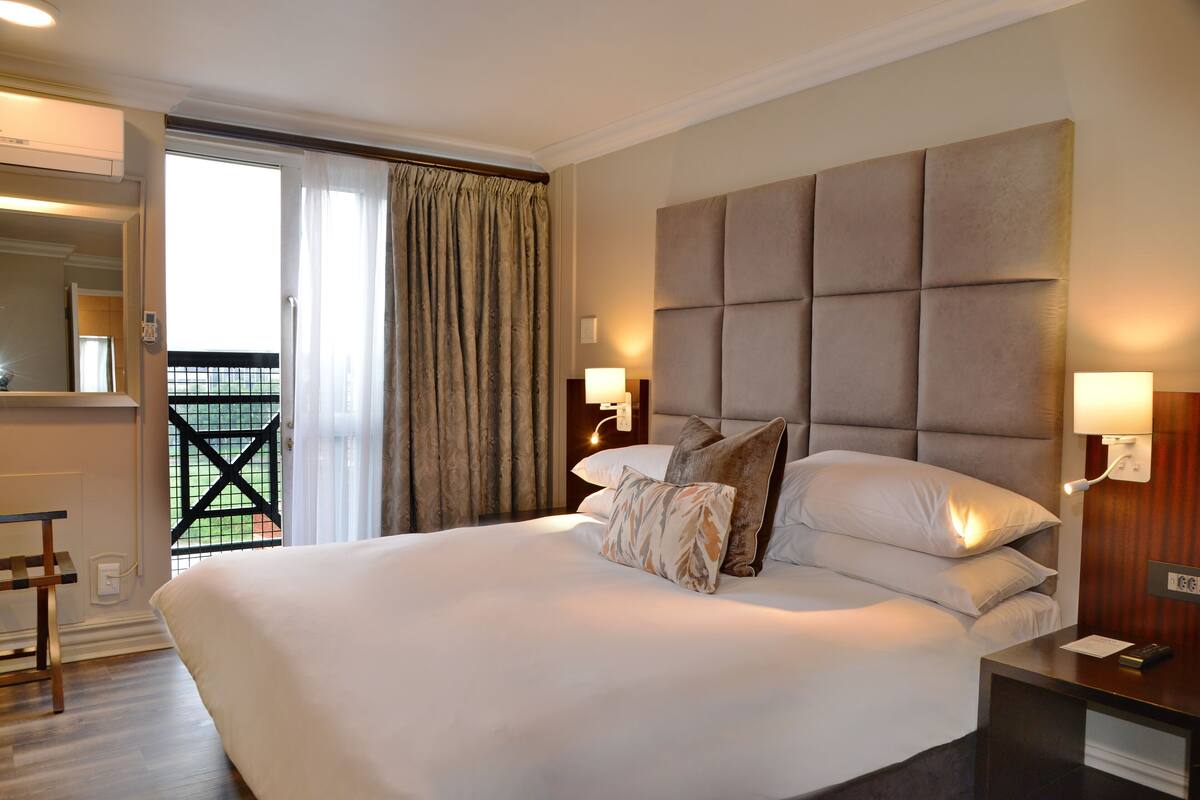 A spacious bedroom with a large bed, a wooden nightstand, a mirror, and a view of a balcony.