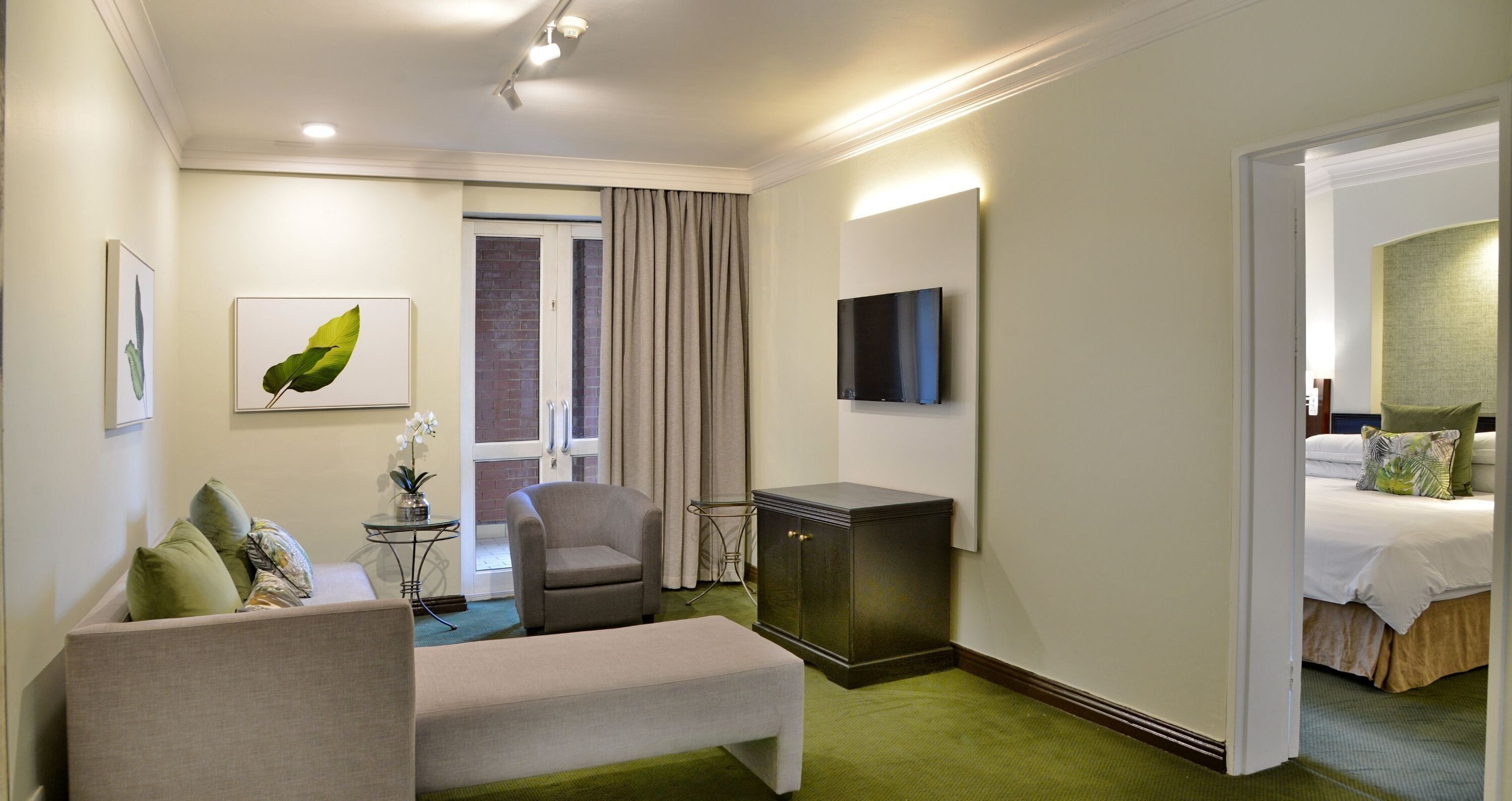 A hotel room with a bed, a seating area with a sofa and armchair, a small table with a plant, a TV mounted on the wall, and a door leading to another room.