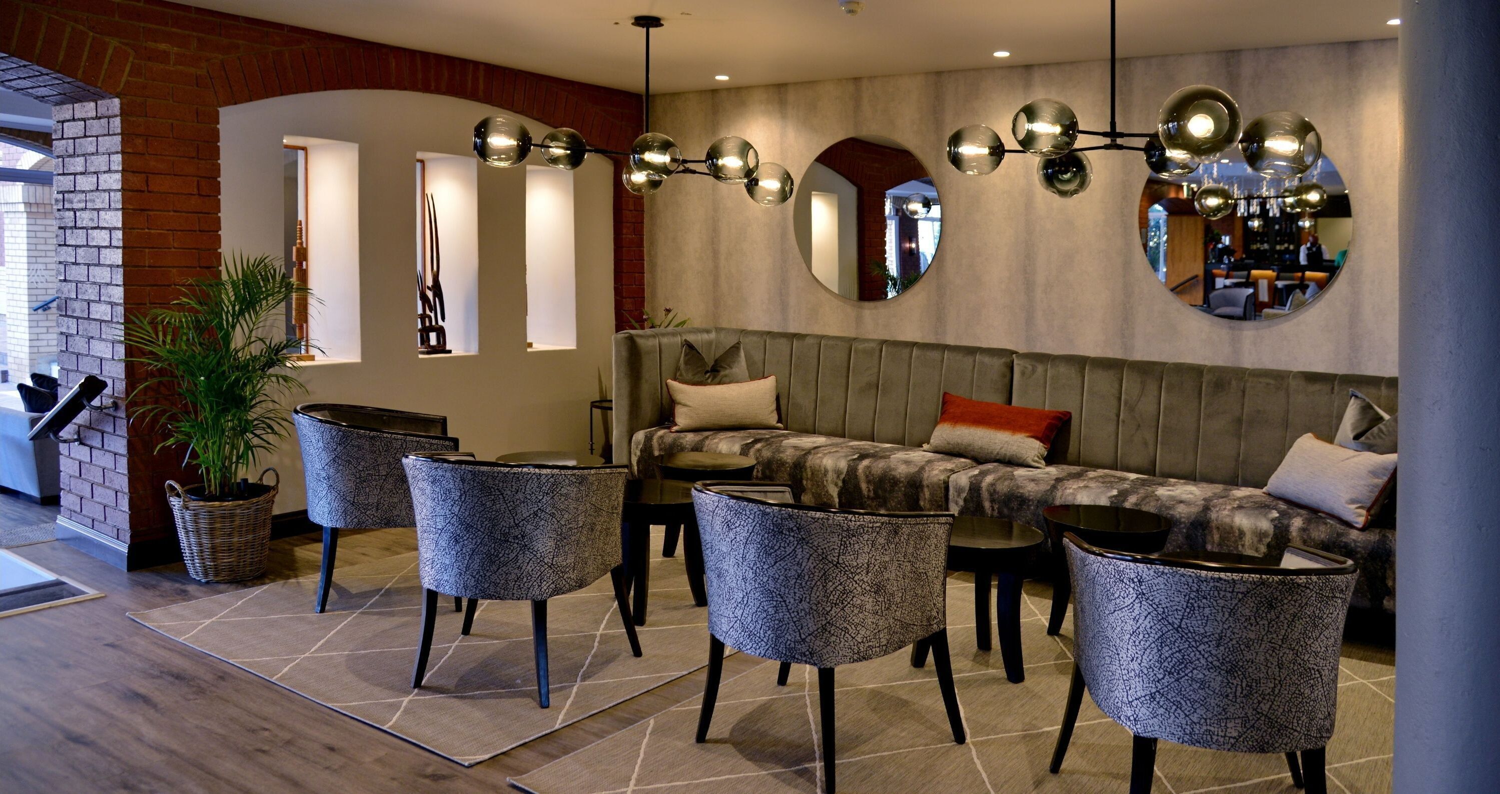 A modern lounge area with a sofa, armchairs, and a dining table. There is a large mirror on the wall and a potted plant.