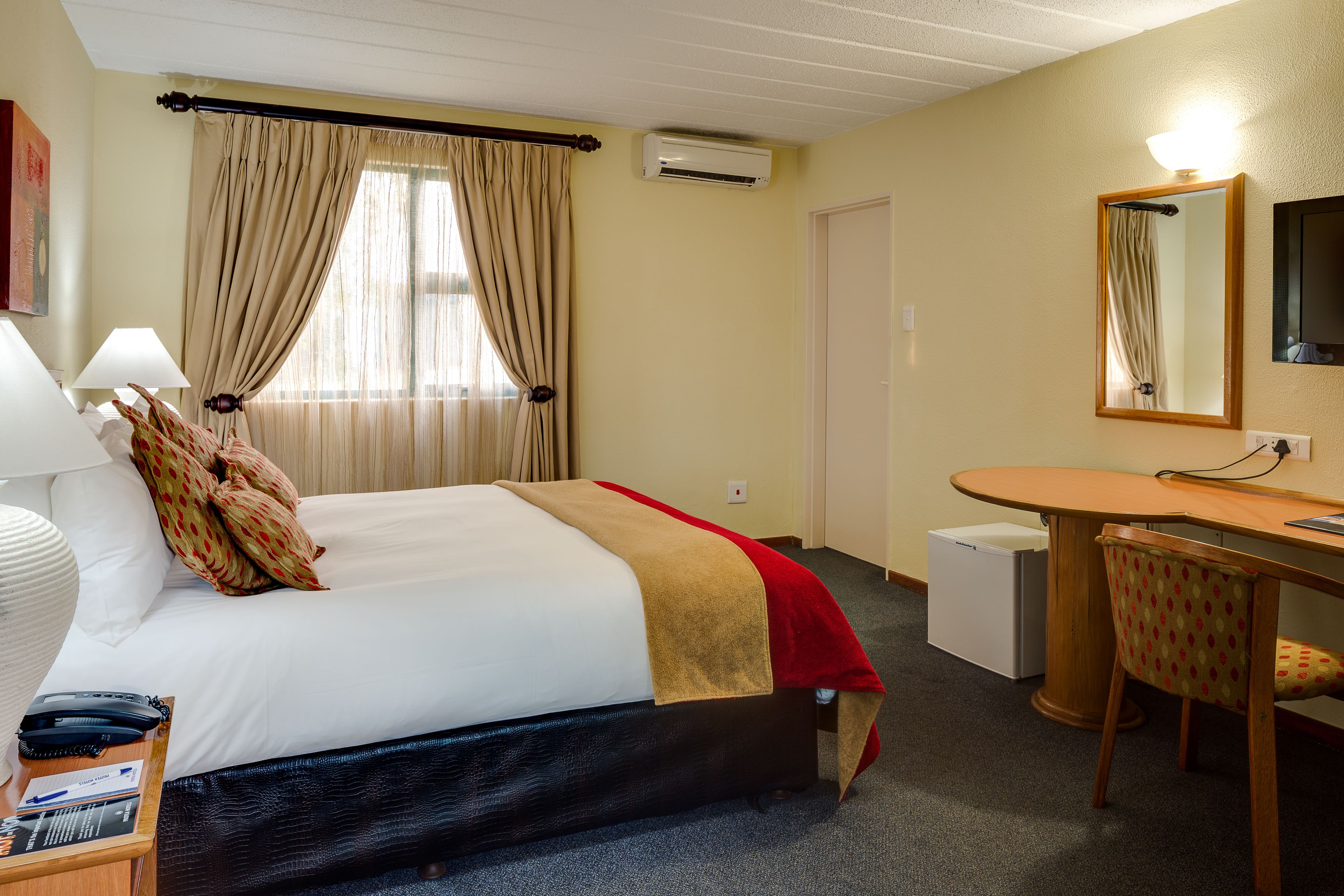 A hotel room with a bed, a desk, a chair, a lamp, a mirror, and an air conditioning unit.