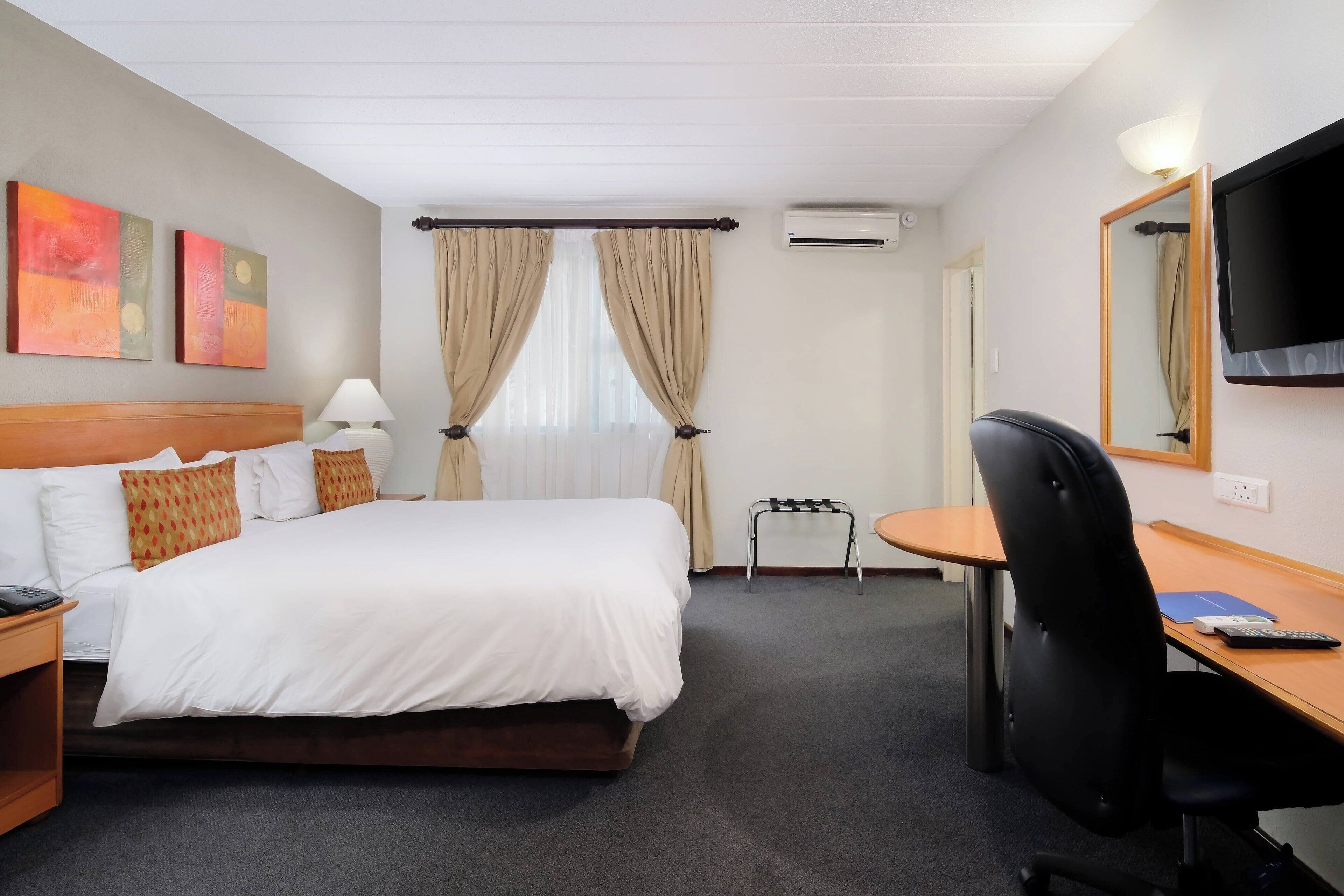 A hotel room with a bed, a desk with a chair, a TV, and an air conditioning unit.