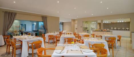 A dining area with multiple tables set for guests, featuring white tablecloths and wooden chairs.