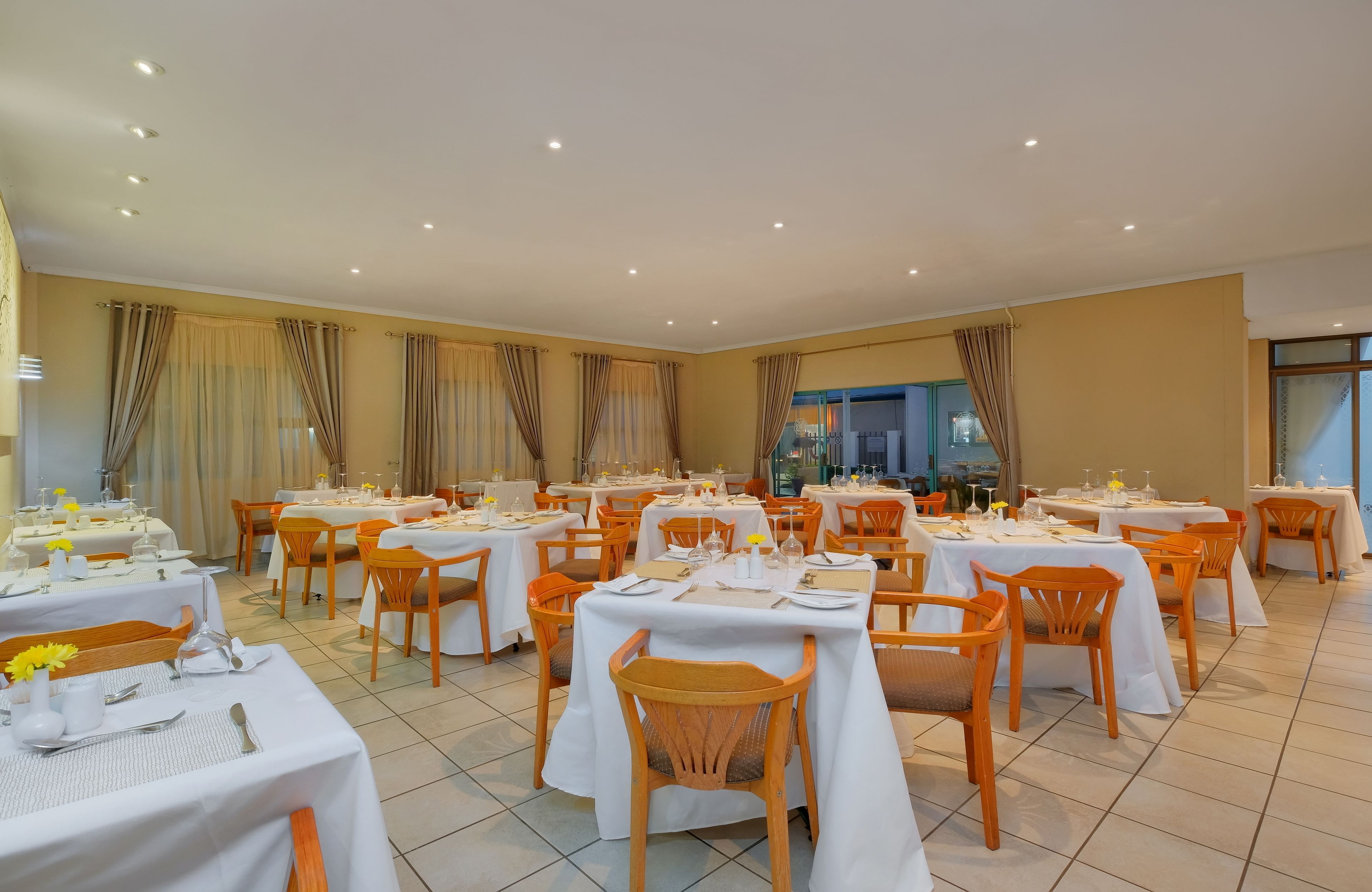Comedor con varias mesas dispuestas para el servicio, sillas de madera y vista a la cocina por un puerta de vidrio.