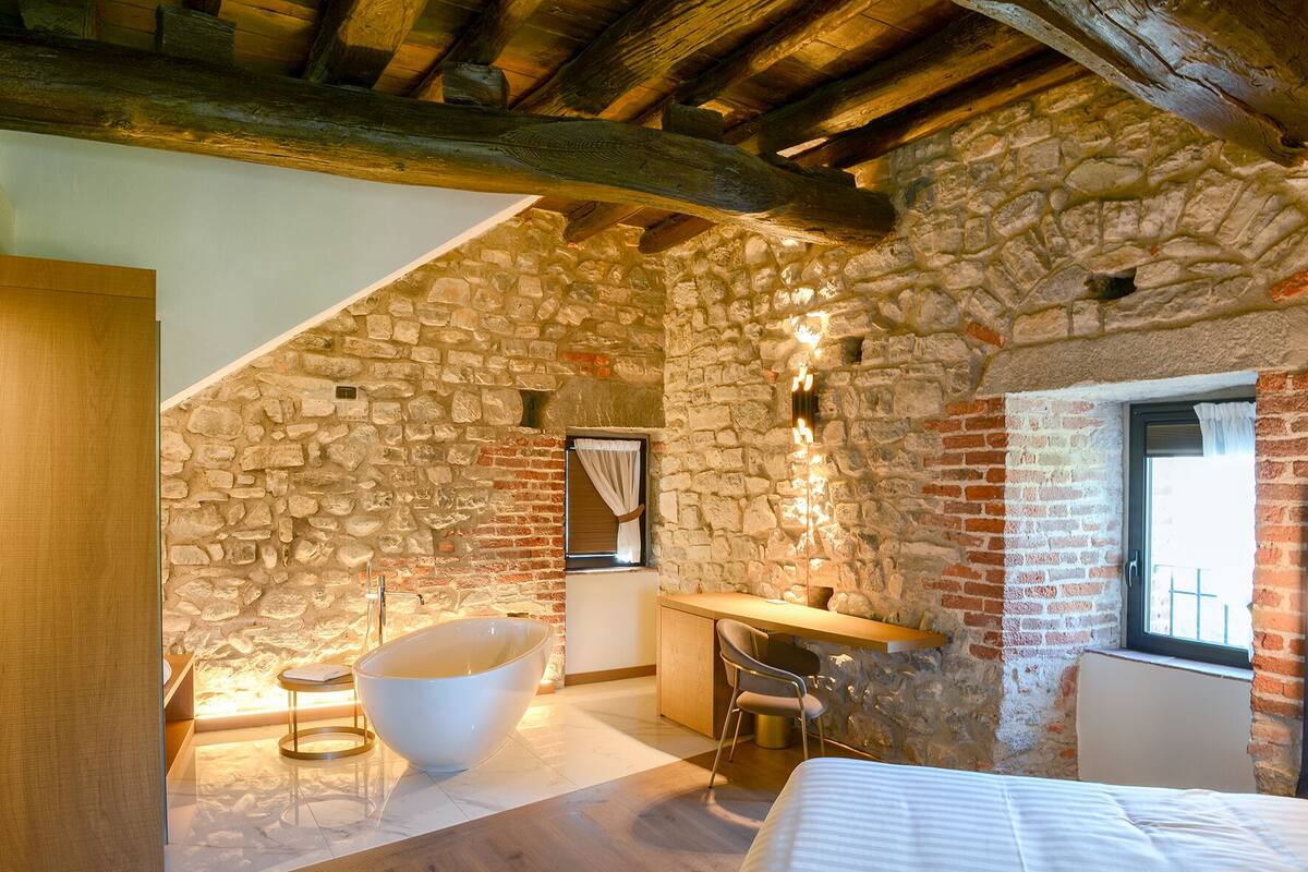 A bedroom with a stone wall, a bathtub, a wooden ceiling, and a window.
