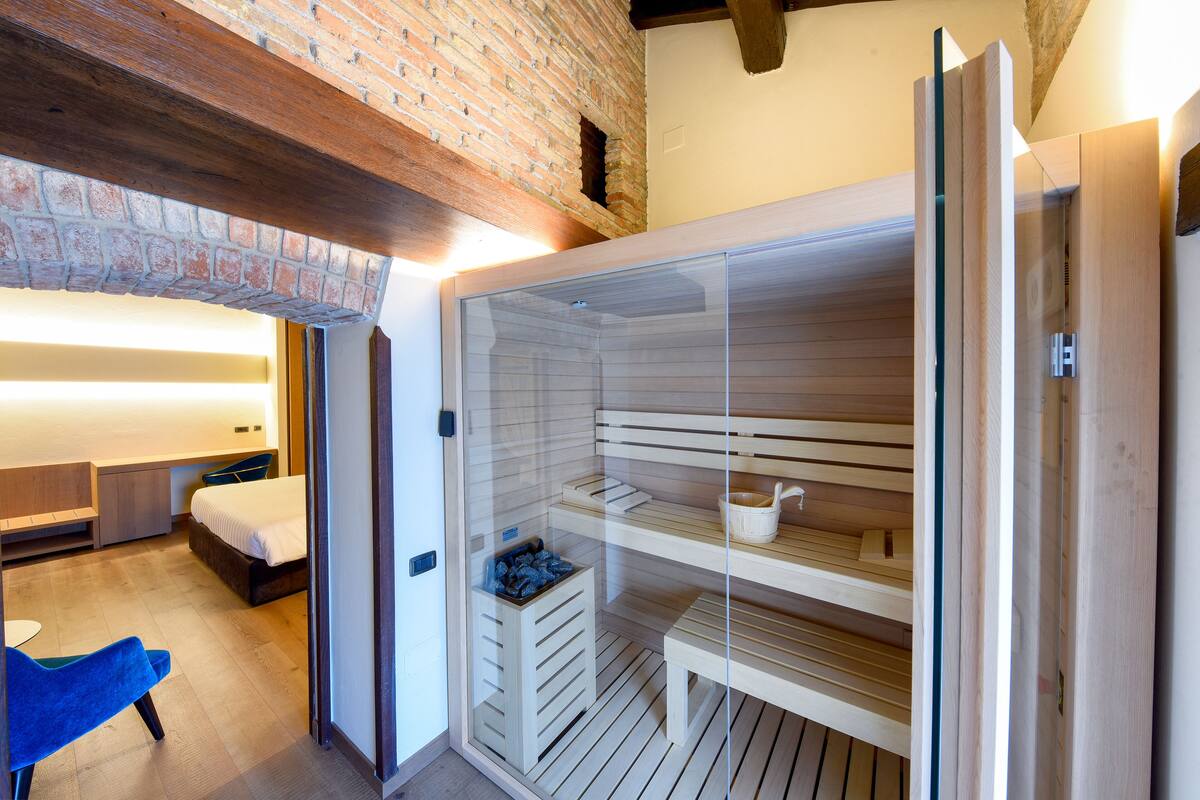 A modern hotel room with a glass-enclosed sauna, wooden flooring, a bed, a desk, and a blue chair.