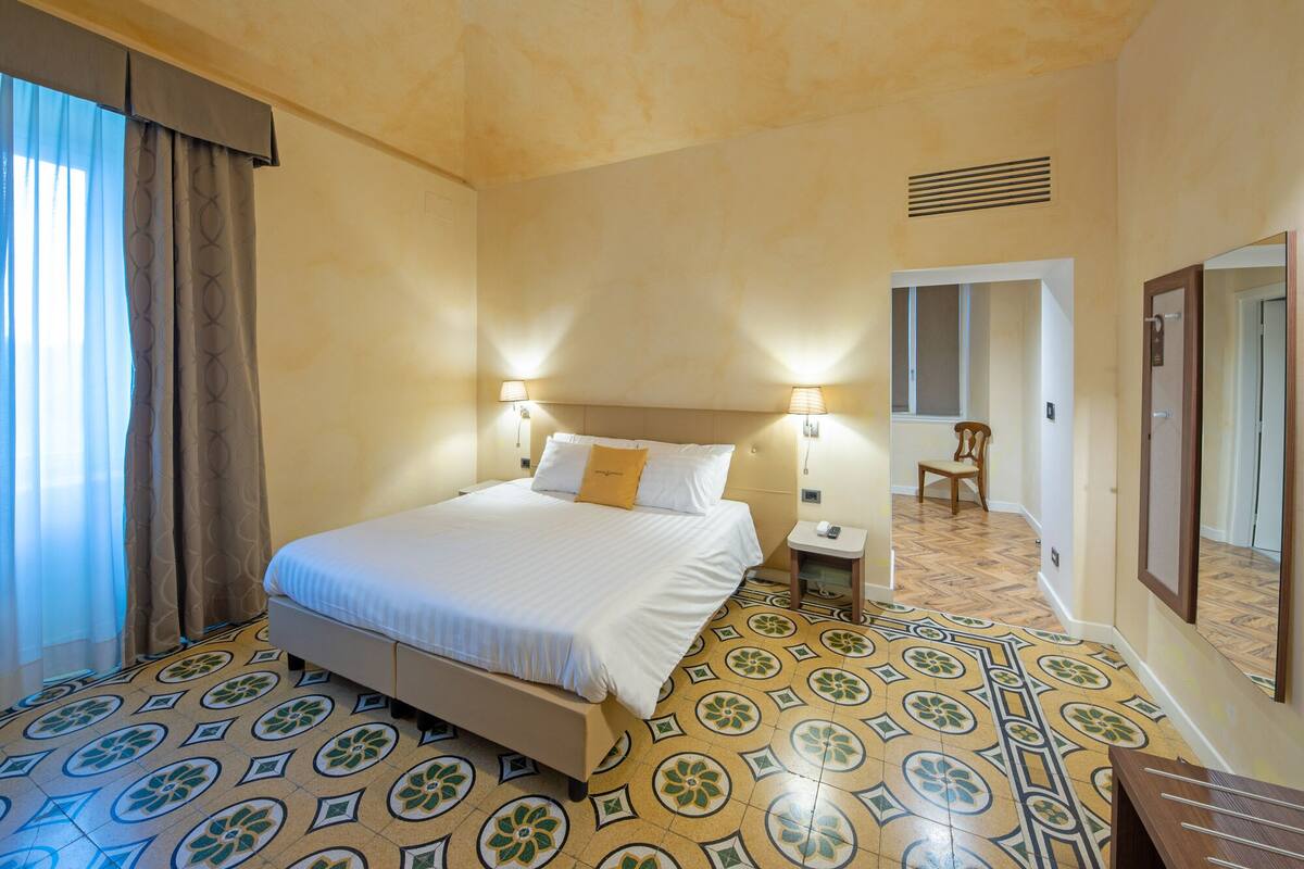 A hotel room with a bed, two bedside lamps, a chair, a mirror, and a patterned floor.