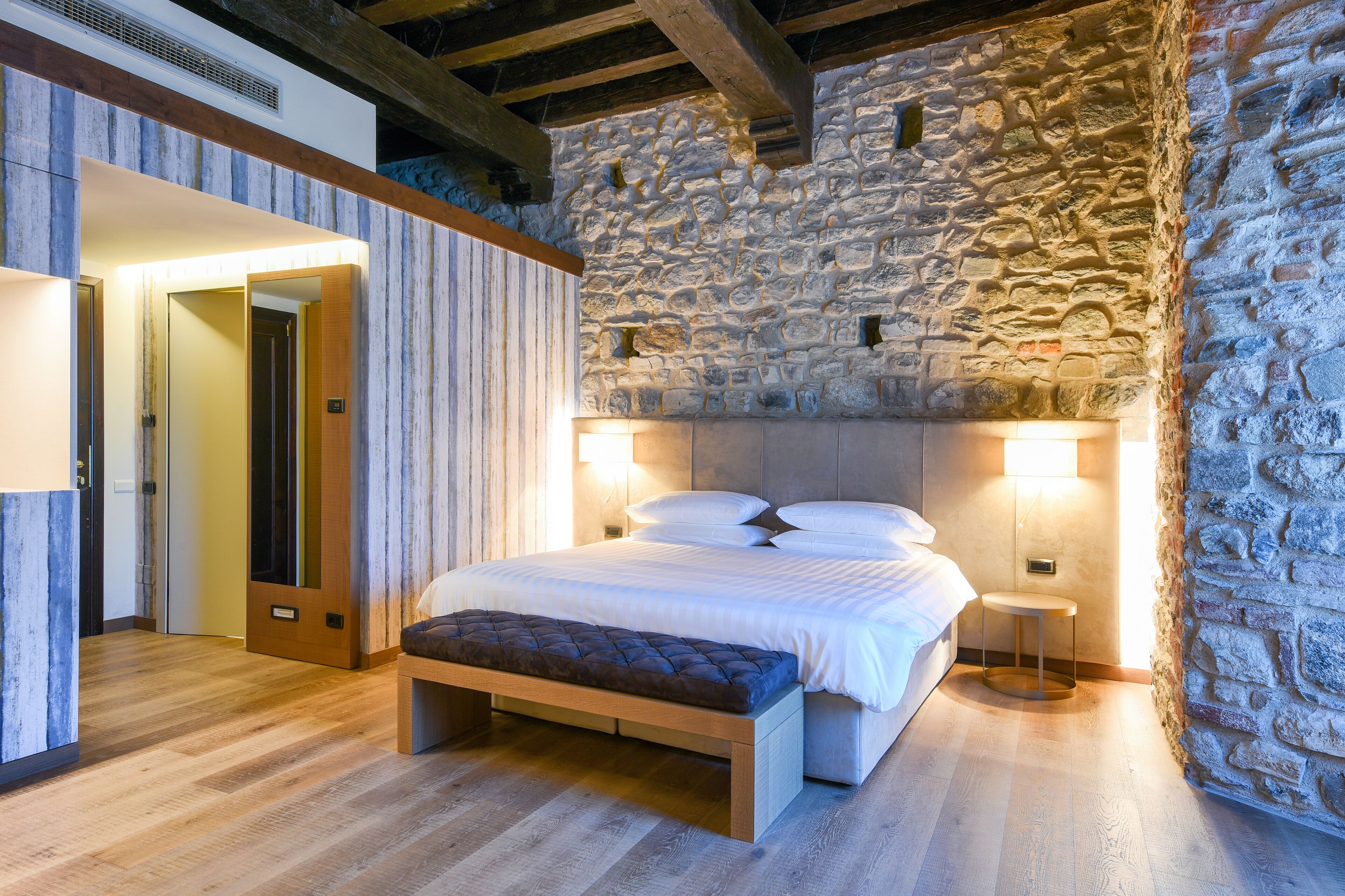 Una camera da letto con parete in pietra, una panca di legno, un letto con biancheria bianca e due lampade ai comodini.