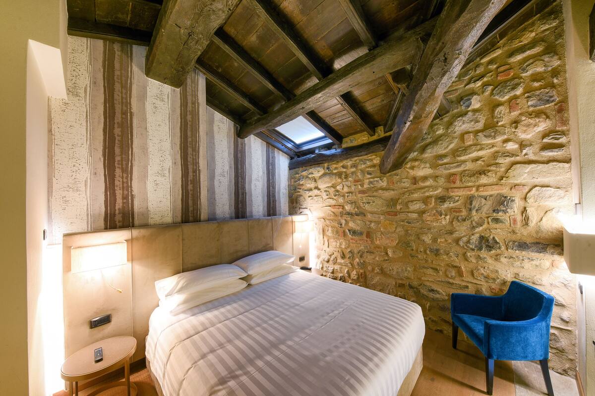 A bedroom with a stone wall, a wooden ceiling, a bed with white linens, a small round table, and a blue chair.